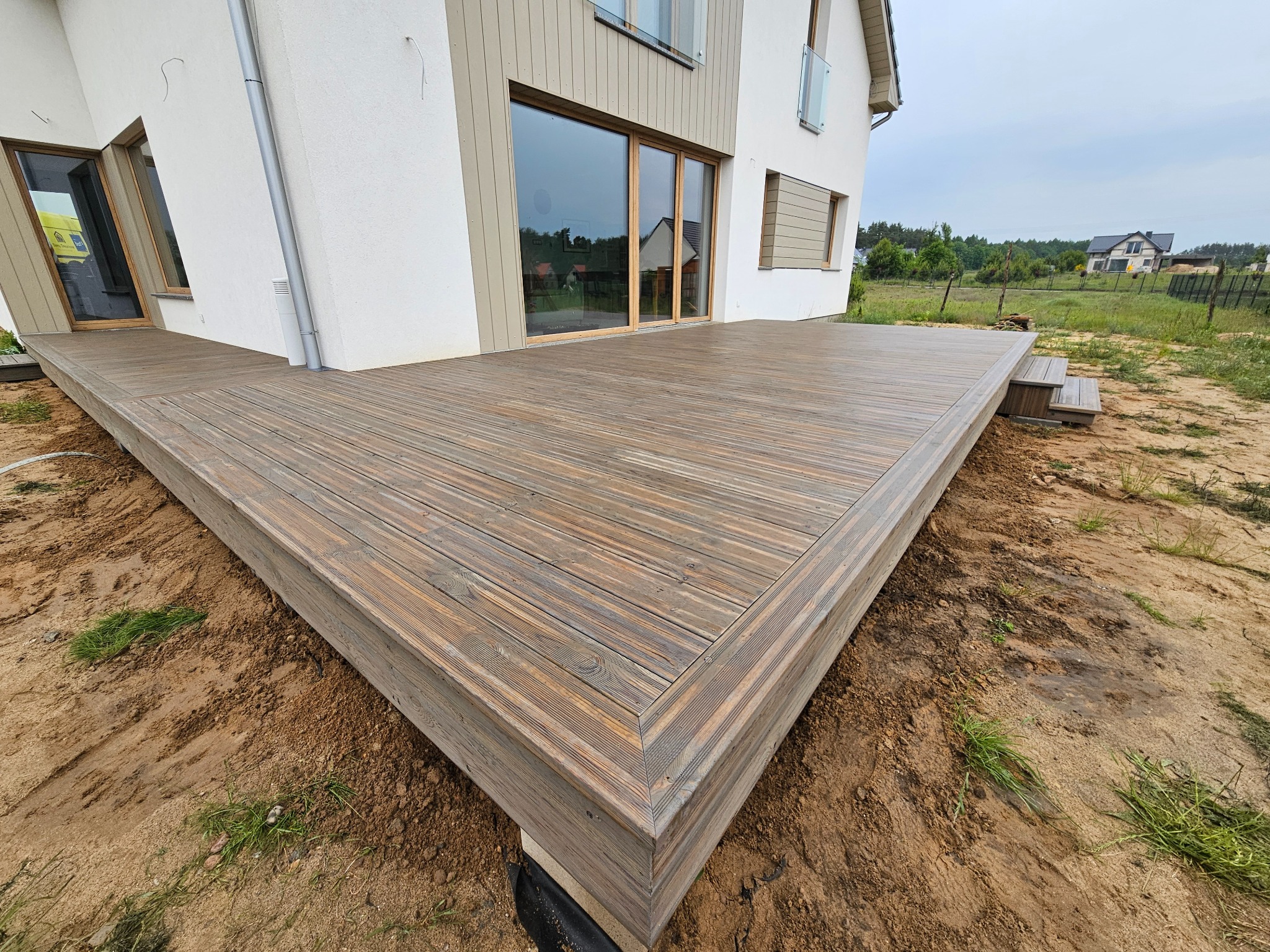 Drewniany taras przylegający do nowoczesnego domu szkieletowego z dużymi oknami, widok z niskiej perspektywy, ukazujący detale desek i połączenia na rogu tarasu.