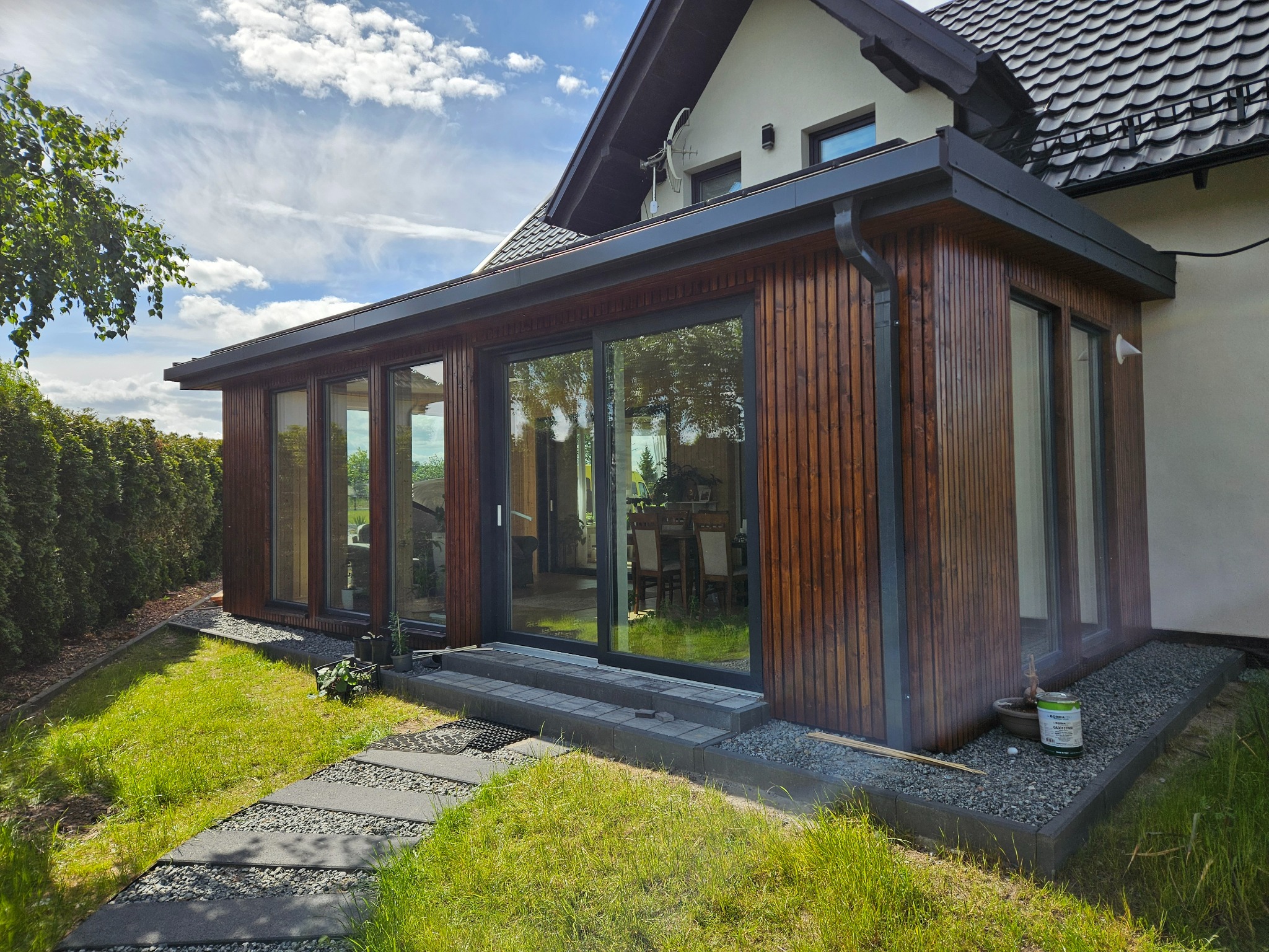 Nowoczesna dobudówka z drewna w domu jednorodzinnym, z dużymi przesuwnymi drzwiami i oknami, otoczona zielonym trawnikiem i ścieżką z płyt.