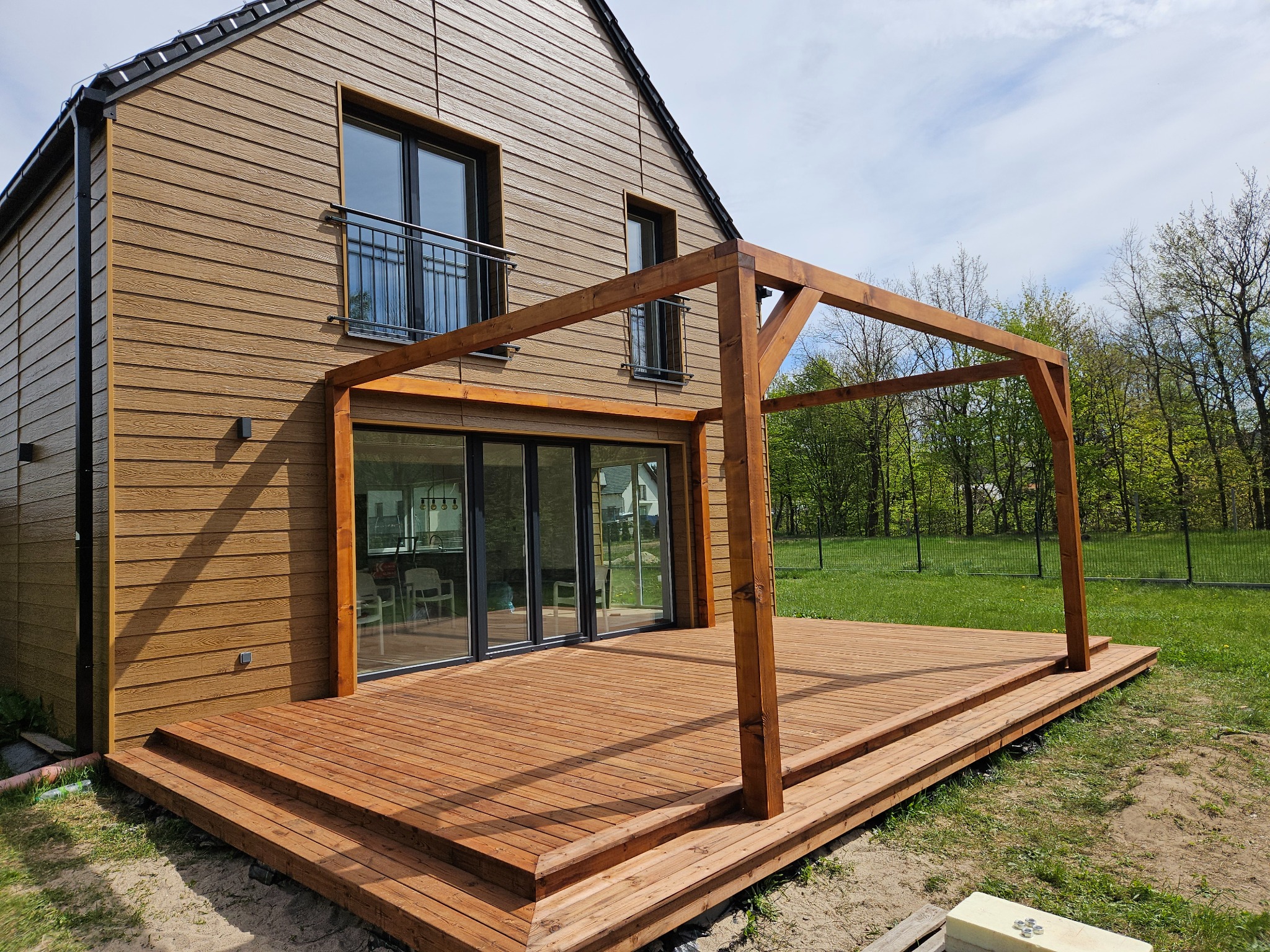 Drewniany taras z pergolą przy brązowym domu szkieletowym z oknami balkonowymi i dużymi przesuwnymi drzwiami. Widok na zieleń w tle.
