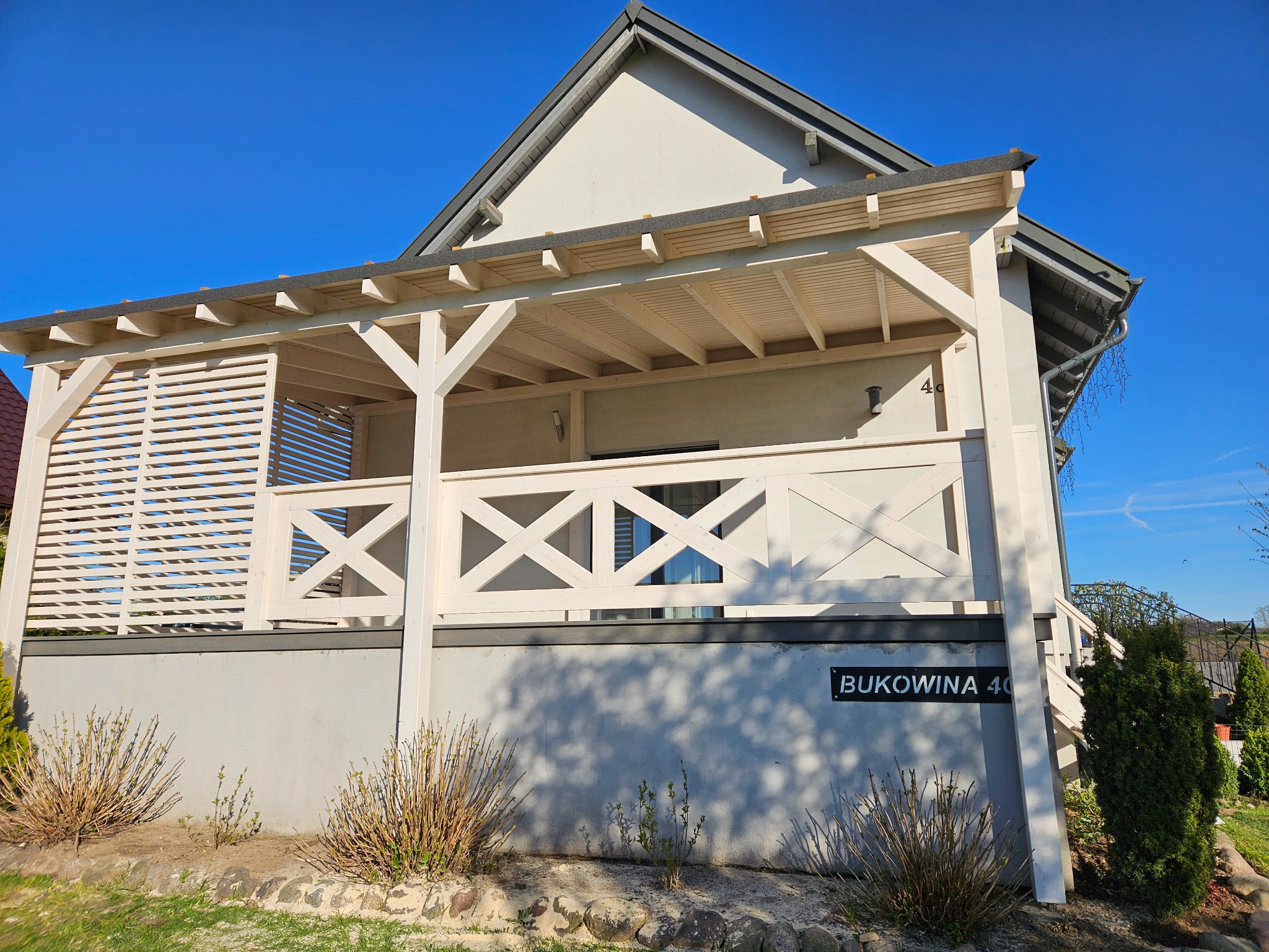 Jasny dom z drewnianą pergolą i ażurową balustradą, Bukowina 40 na elewacji, błękitne niebo w tle. Nowoczesna architektura z elementami drewna.