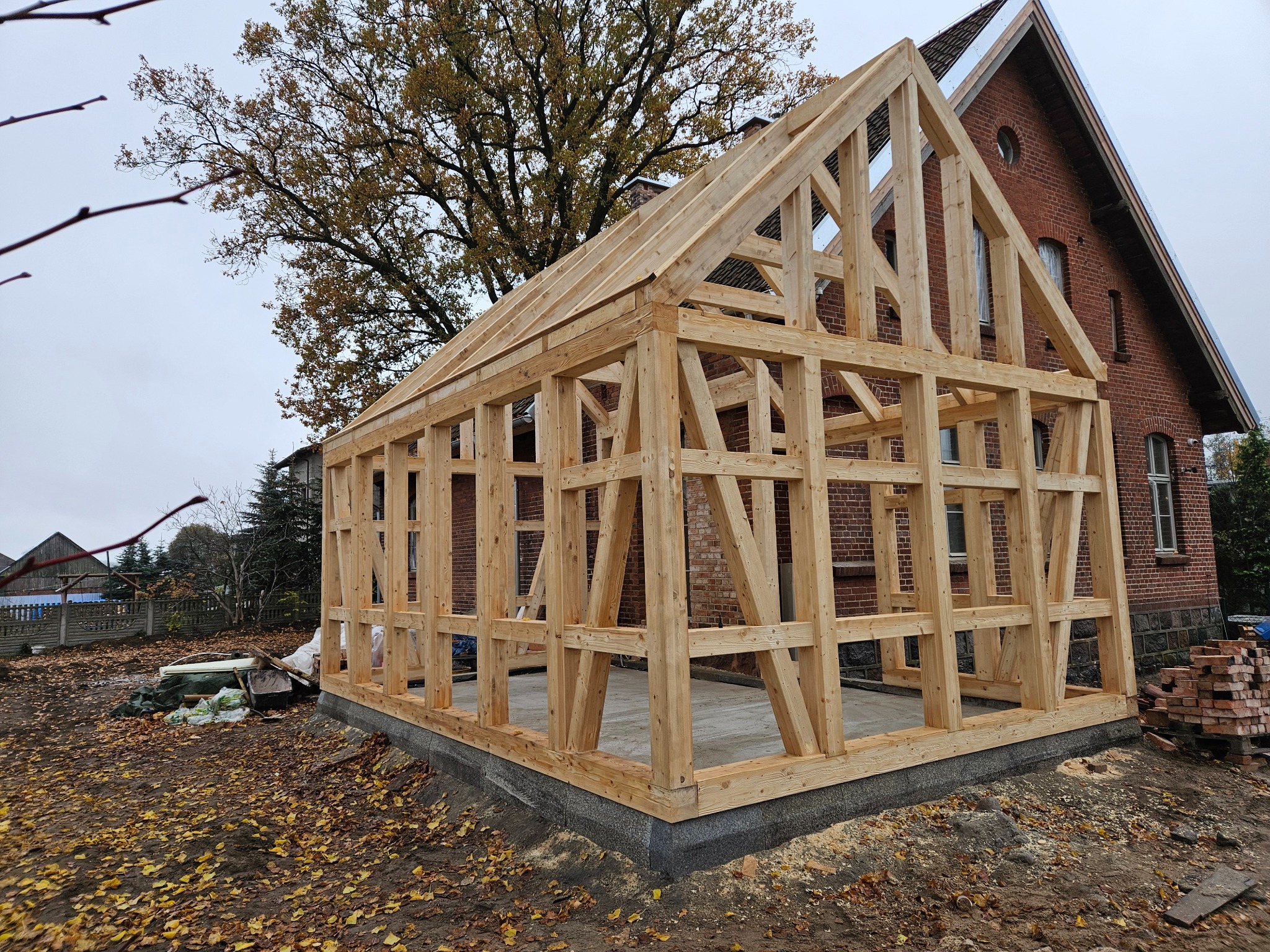 Konstrukcja domu szkieletowego w trakcie budowy, widoczny drewniany szkielet na fundamencie, w tle budynek z czerwonej cegły i drzewo.