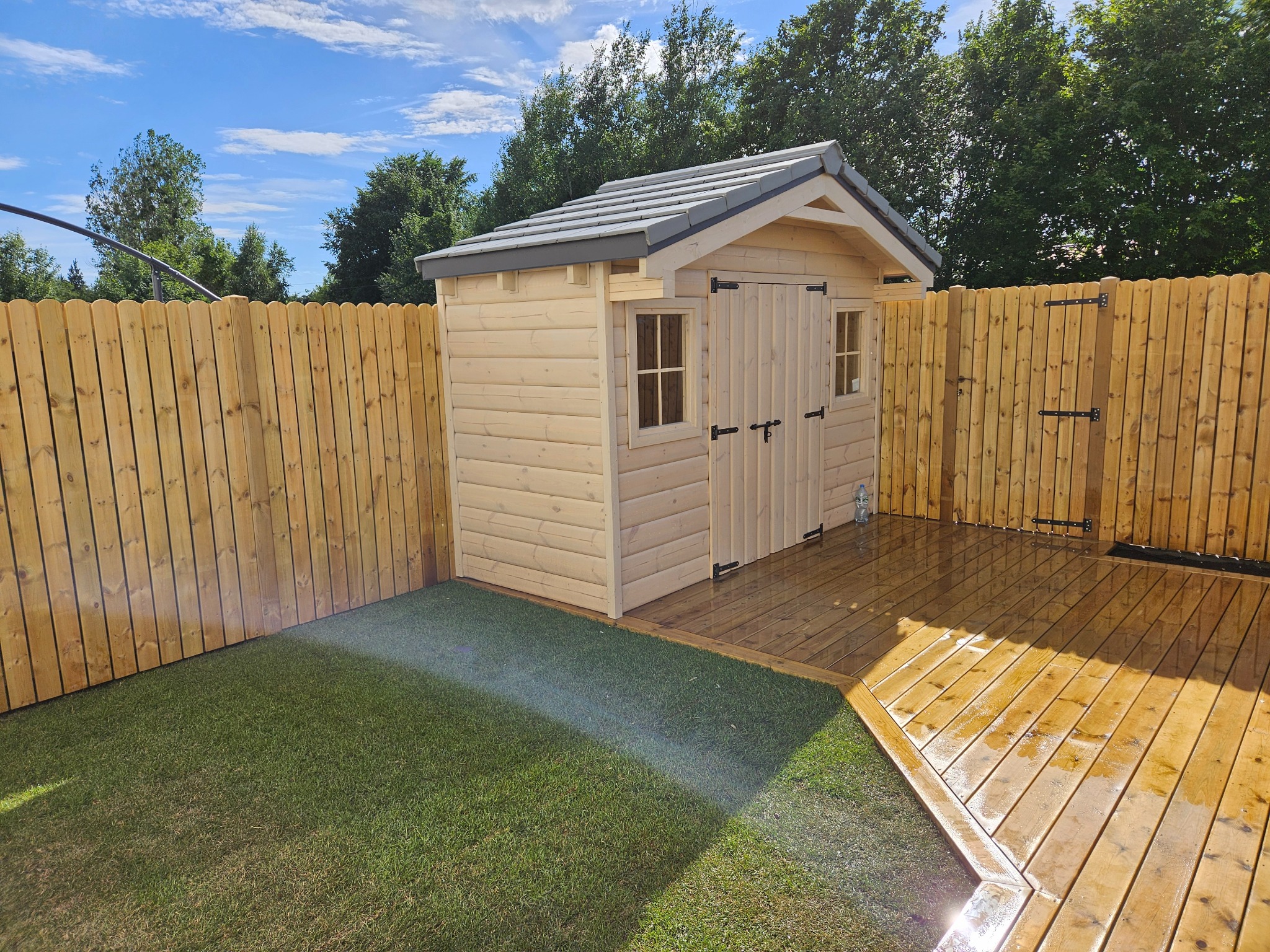 Drewniany domek narzędziowy z szarą dachówką, otoczony drewnianym płotem i sztuczną trawą, przylegający do drewnianego tarasu w słoneczny dzień.