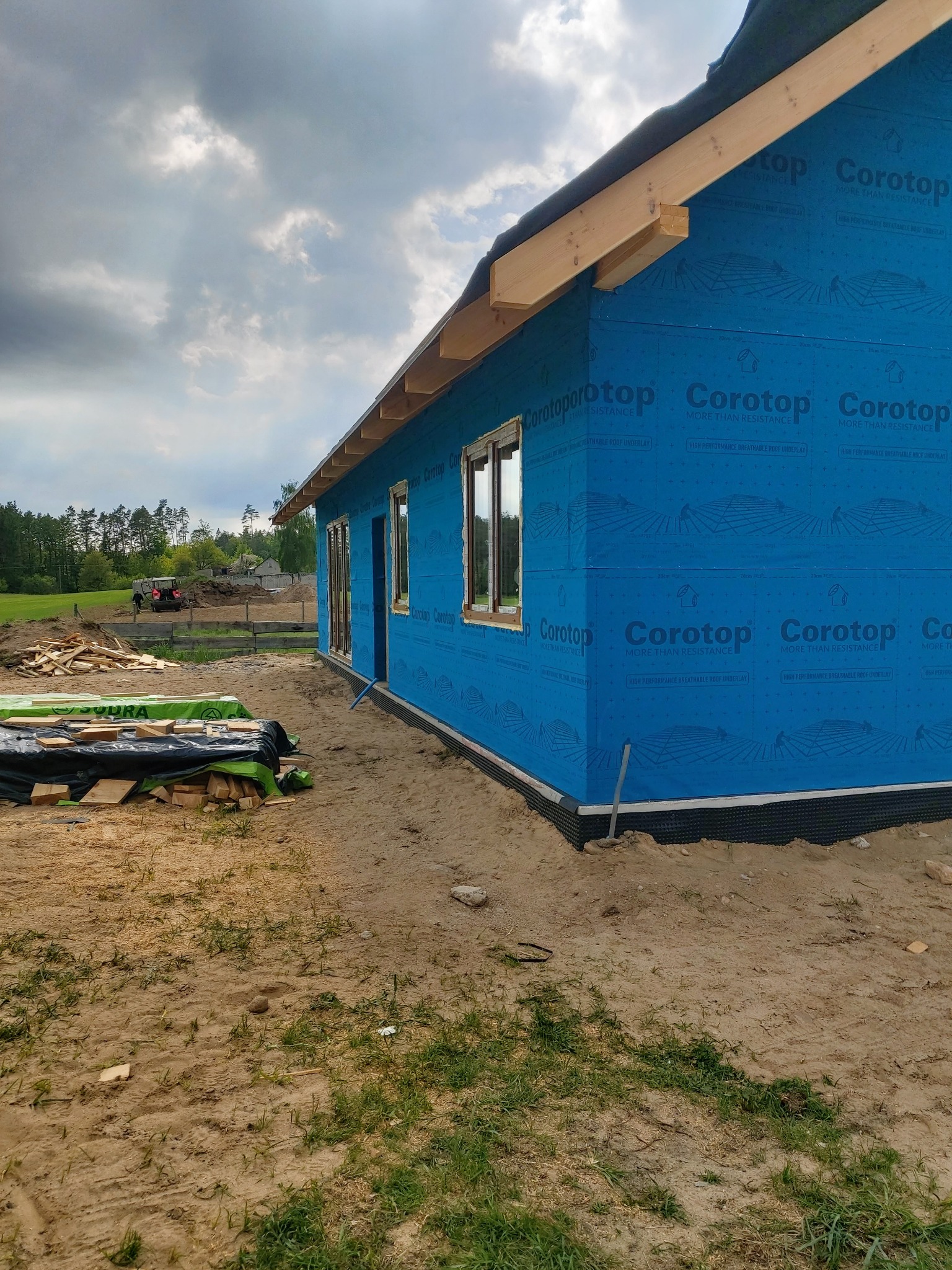 Budowa domu szkieletowego w trakcie: ściany pokryte niebieską membraną Corotop, widoczne drewniane elementy konstrukcyjne, teren budowy z materiałami.