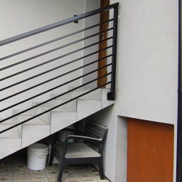 KriStalSpaw - Zewnętrzne schody z czarną, metalową balustradą o poziomych prętach. Białe płytki schodowe, krzesło i wiadro pod schodami. Ściana z rurą spustową.