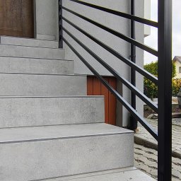 KriStalSpaw - Zewnętrzne schody z szarymi płytkami i czarną, nowoczesną balustradą z poziomych profili. Widoczny fragment drzwi wejściowych i otoczenia budynku.