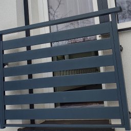 KriStalSpaw - Szary, poziomy panel balustrady balkonowej, zamontowany na białej elewacji budynku, widoczny przez okno z refleksami drzew.