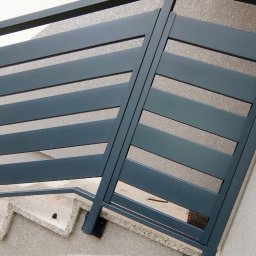 KriStalSpaw - Nowoczesna, pozioma brama wjazdowa z szarych, metalowych paneli, zamontowana na schodach wejściowych. Widok z dołu, podkreślający kąt nachylenia i minimalistyczny design.