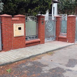 KriStalSpaw - Metalowa brama wjazdowa z furtką, osadzona w murze z czerwonej cegły. Widoczna skrzynka na listy i dekoracyjne elementy metalowe.