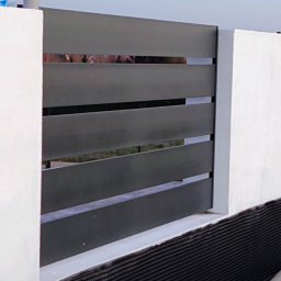 KriStalSpaw - Nowoczesne ogrodzenie z poziomych, grafitowych paneli między białymi słupkami. Widoczne zabezpieczenie fundamentu. Tło stanowią wzgórza i fragment dachu.