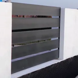 Nowoczesne ogrodzenie z poziomych, grafitowych paneli między białymi słupkami. Widoczne zabezpieczenie fundamentu. Tło stanowią wzgórza i fragment dachu.