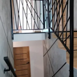 Nowoczesna balustrada schodowa z czarnymi, geometrycznymi elementami na tle betonowej ściany i drewnianych schodów. Unikalny design w loftowym wnętrzu.