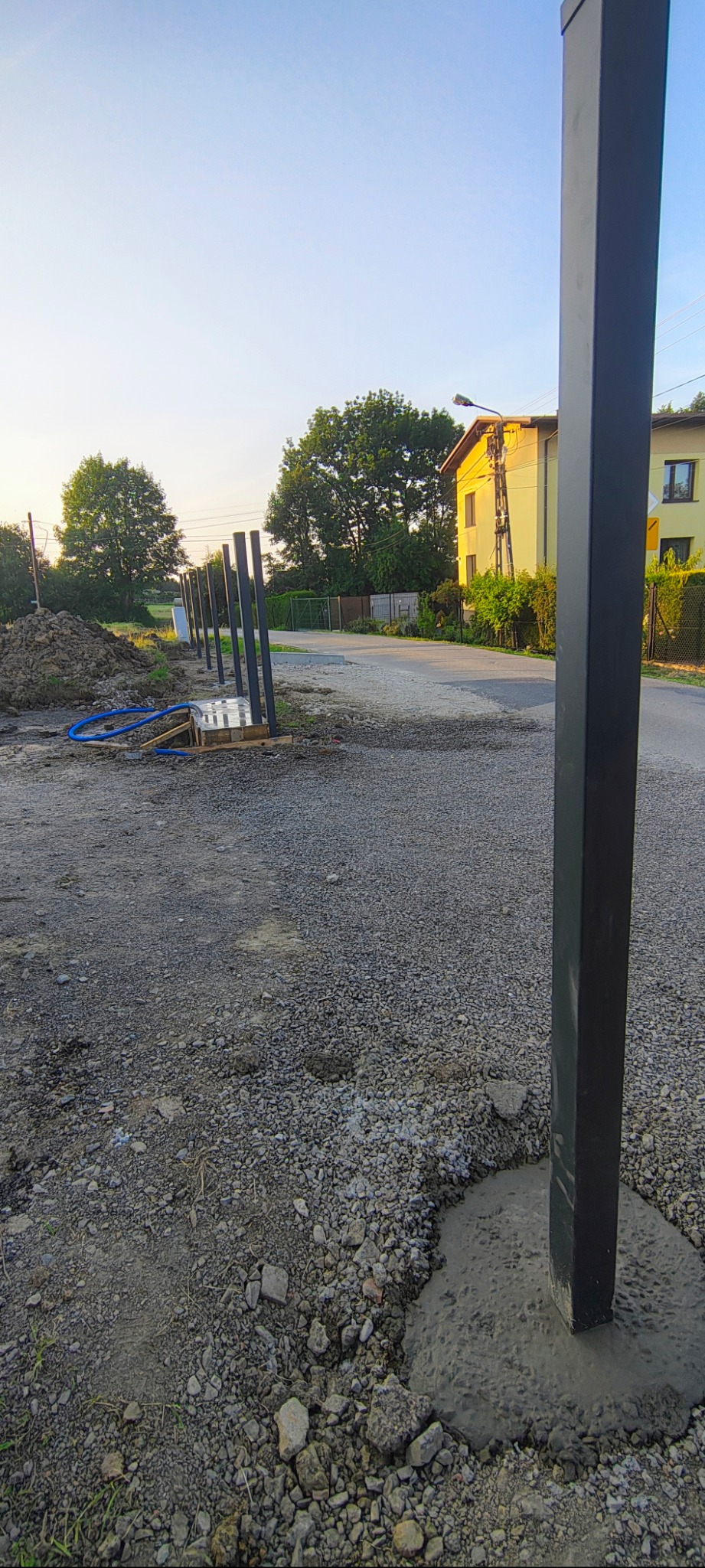 Montaż słupków ogrodzeniowych w ziemi, jeden słupek w betonie na pierwszym planie, rząd słupków w tle, widok na ulicę i domy w oddali.