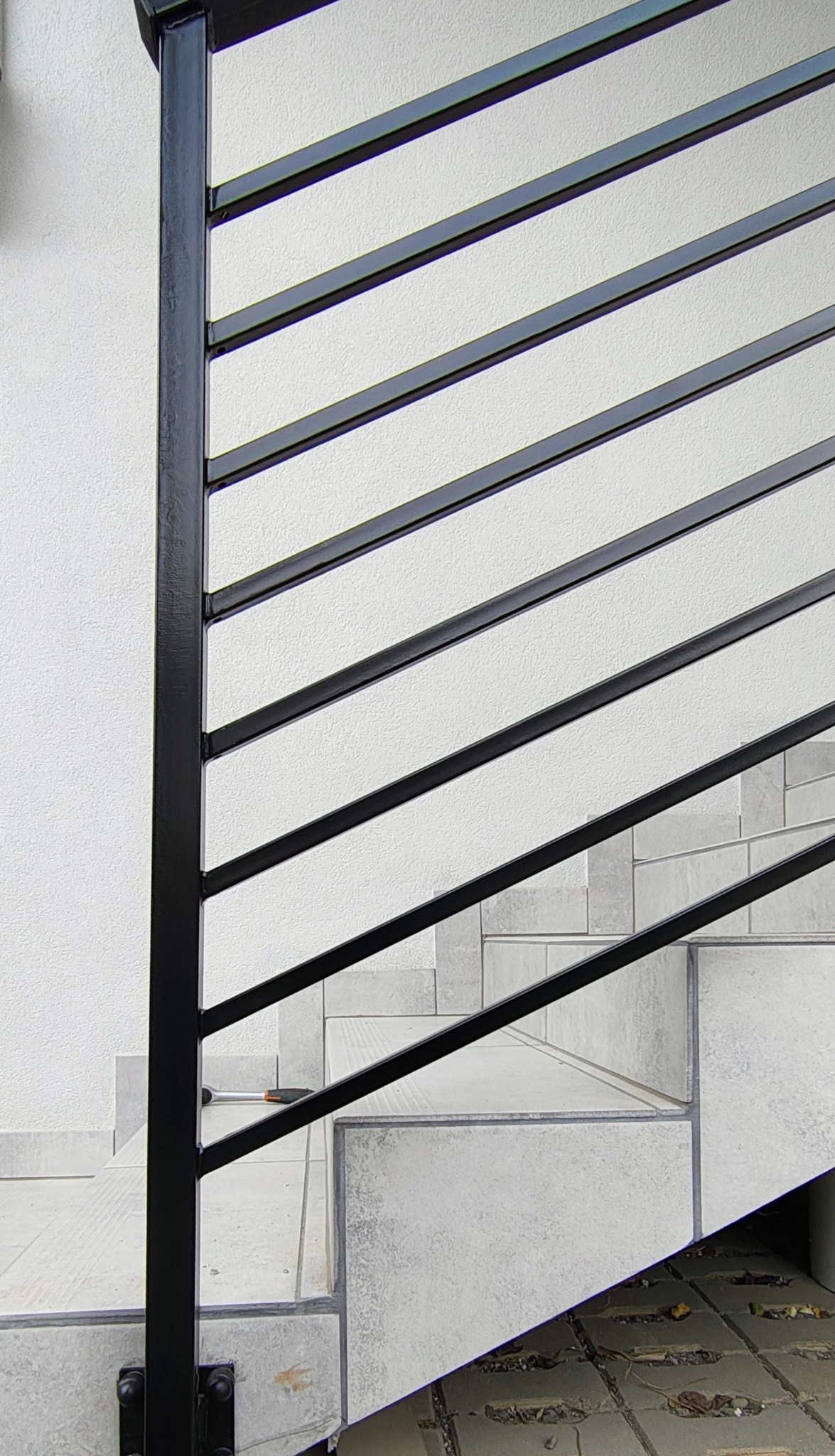 Minimalistyczna balustrada schodowa z czarnymi, poziomymi prętami na tle białej ściany i schodów wyłożonych płytkami. Widoczne mocowania do słupka.