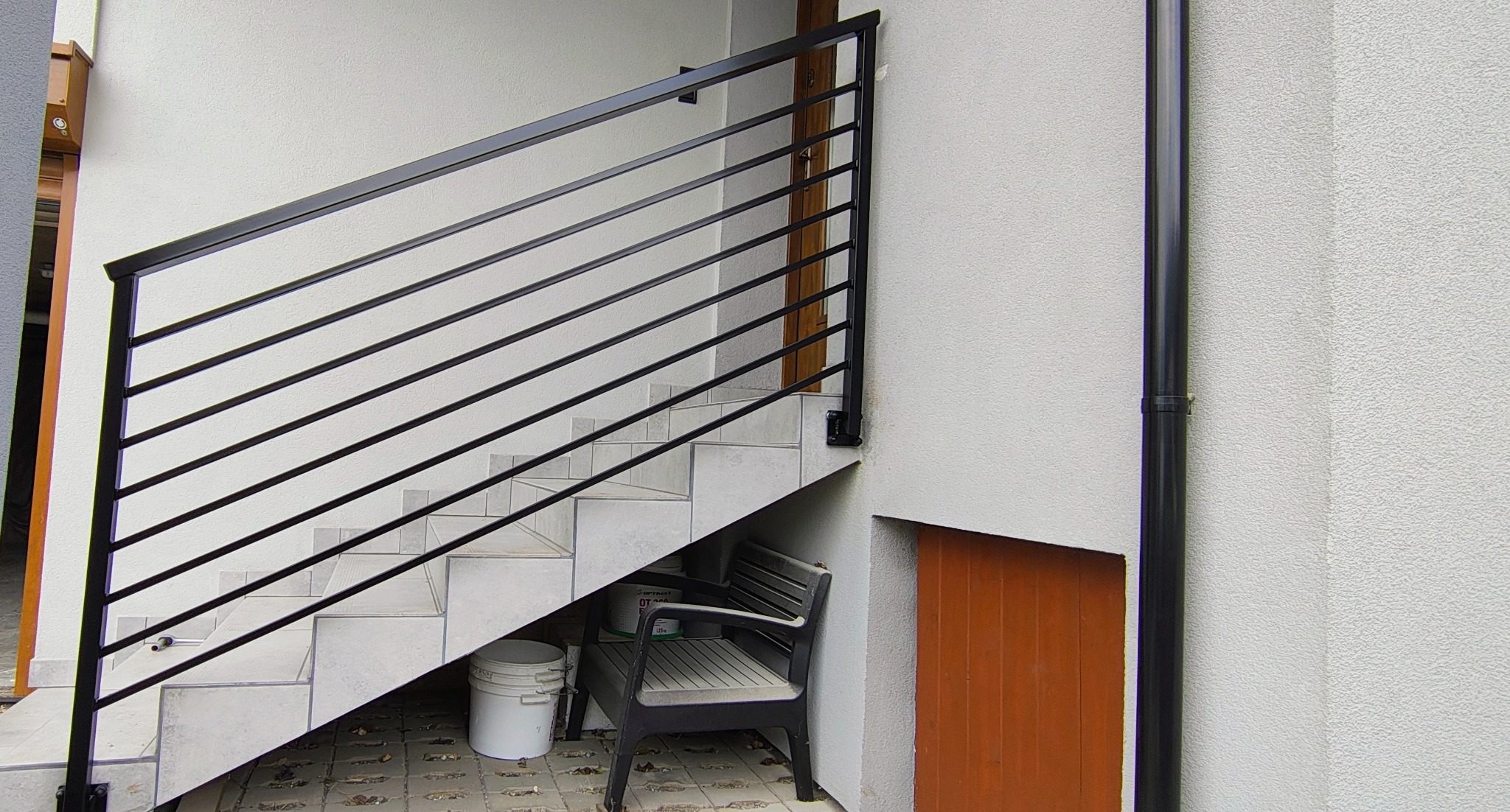 Zewnętrzne schody z czarną, metalową balustradą o poziomych prętach. Białe płytki schodowe, krzesło i wiadro pod schodami. Ściana z rurą spustową.