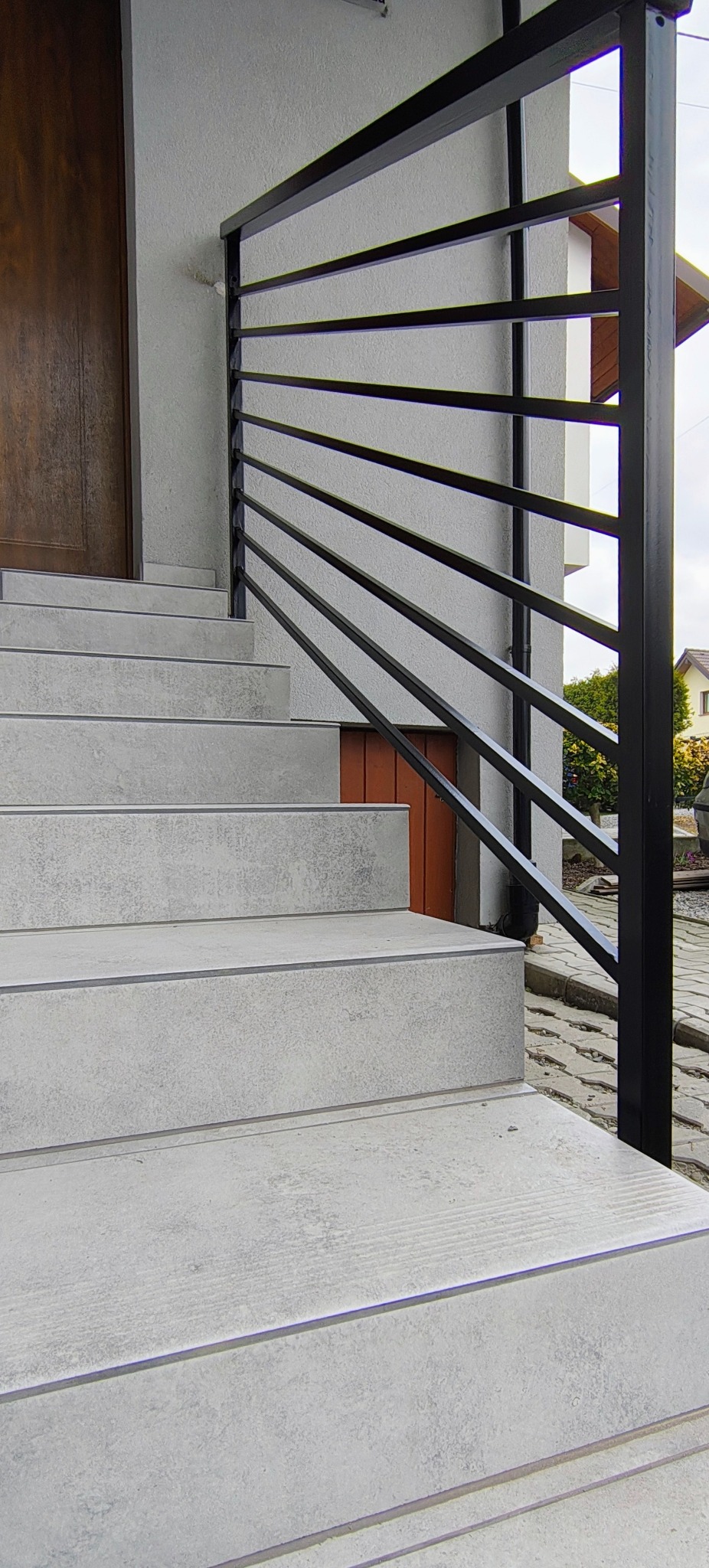 Zewnętrzne schody z szarymi płytkami i czarną, nowoczesną balustradą z poziomych profili. Widoczny fragment drzwi wejściowych i otoczenia budynku.