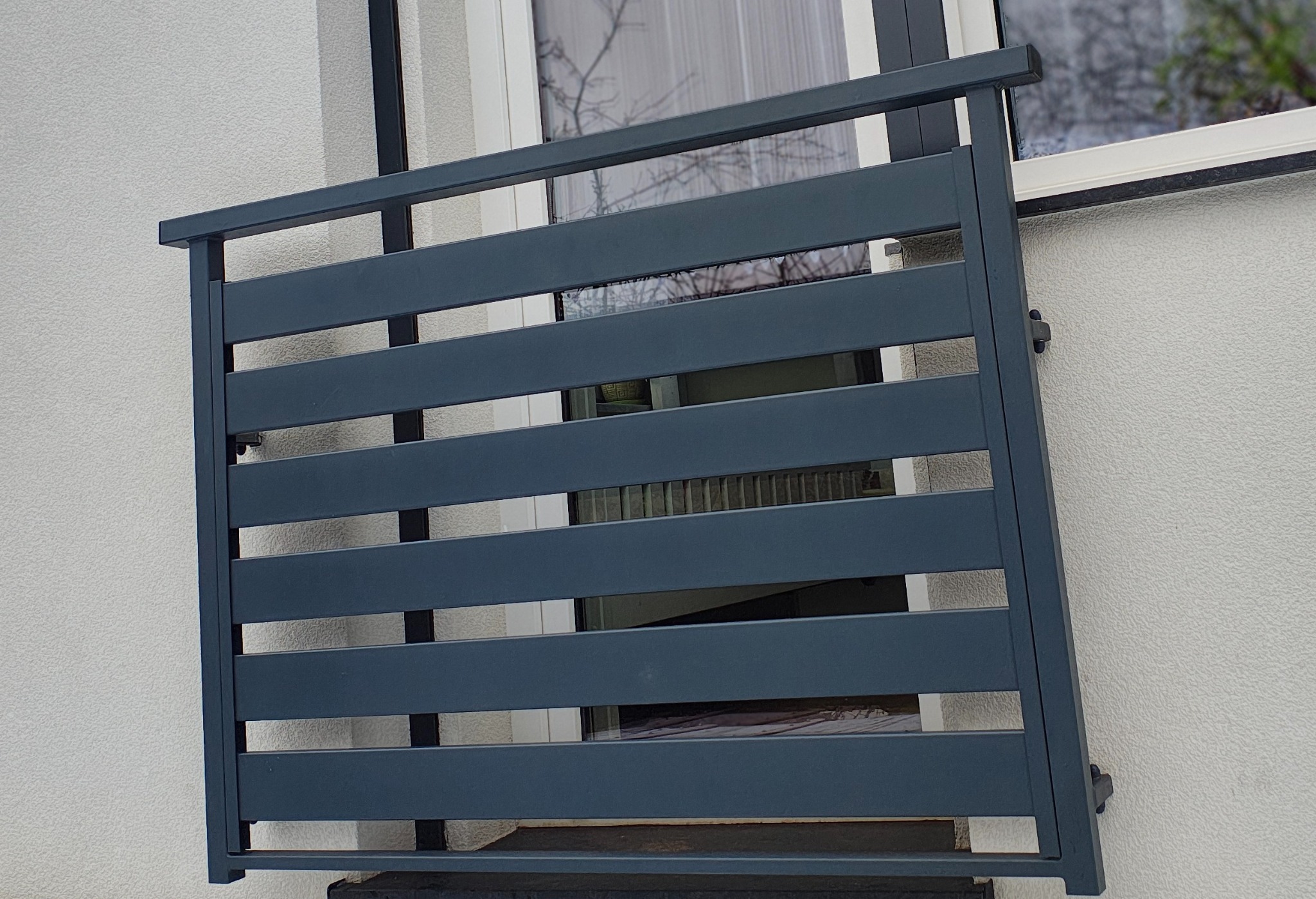 Szary, poziomy panel balustrady balkonowej, zamontowany na białej elewacji budynku, widoczny przez okno z refleksami drzew.