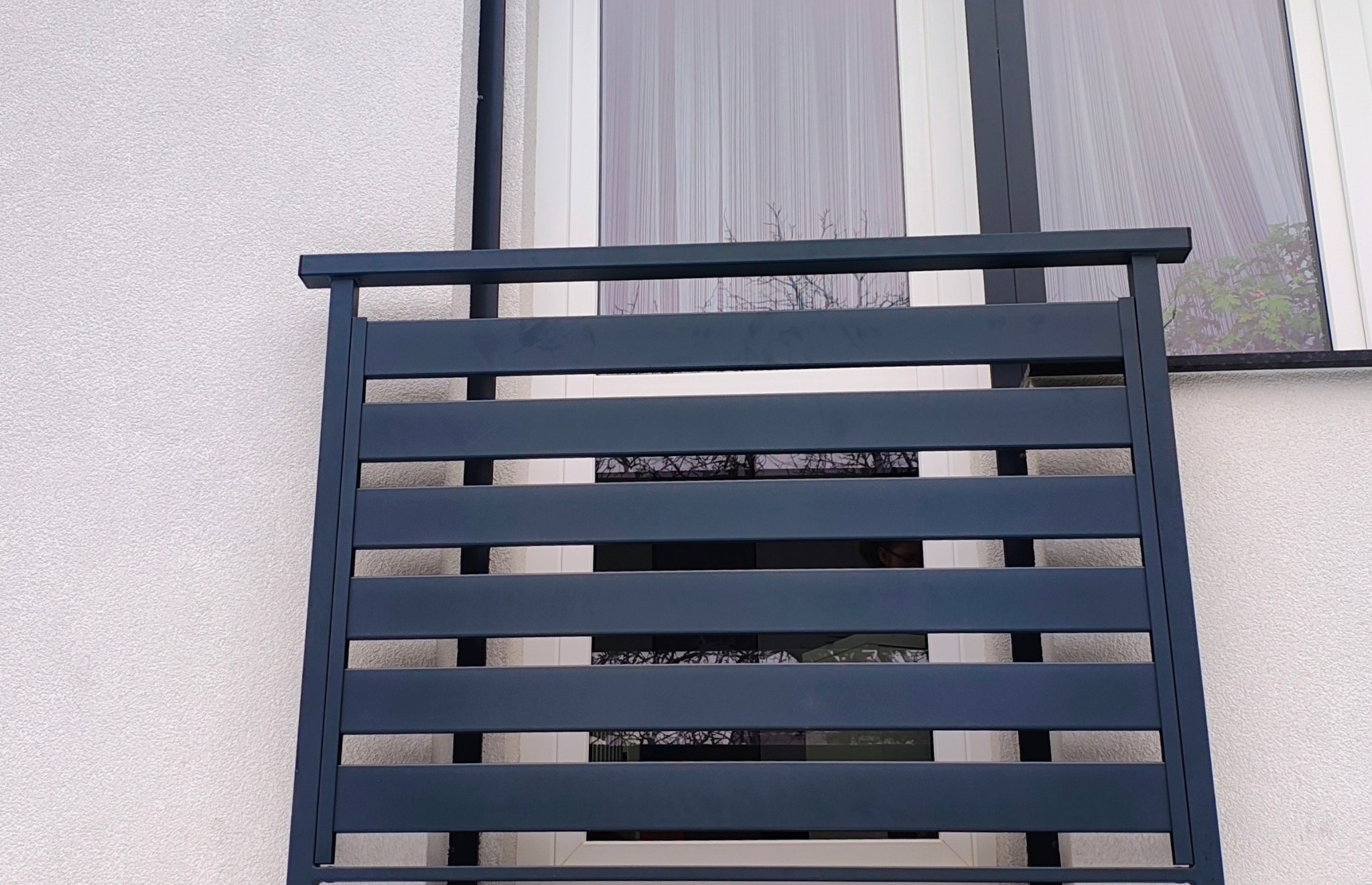 Nowoczesna, grafitowa balustrada balkonowa z poziomych profili, zamontowana na tle białej elewacji i okna z firanką. Minimalistyczny design.
