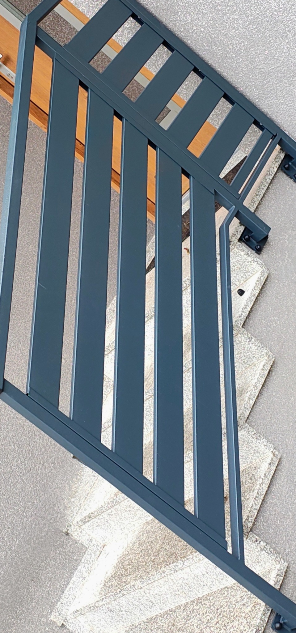 Metalowa balustrada schodowa w odcieniach szarości, z pionowymi szczeblami i poziomymi elementami łączącymi, zamontowana na jasnych, granitowych stopniach.