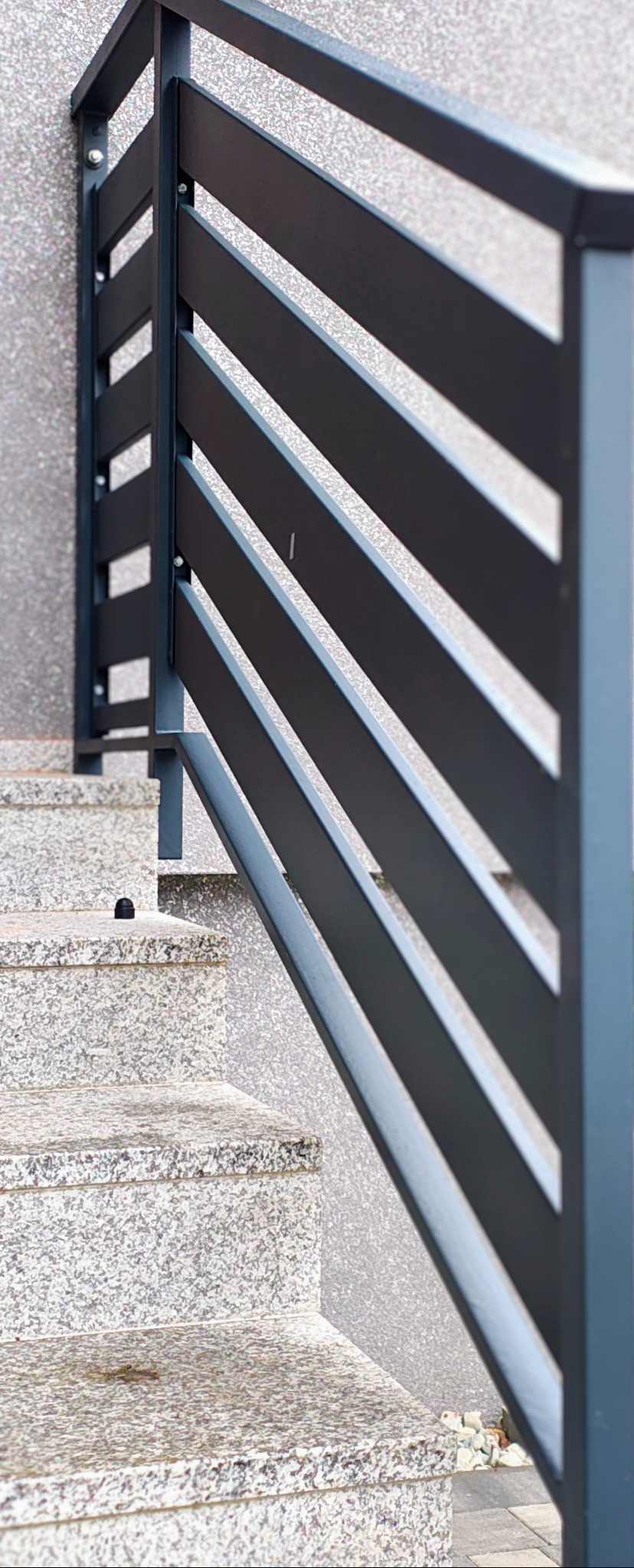 Nowoczesna balustrada metalowa w kolorze antracytu, z poziomymi szczeblami, zamontowana przy schodach zewnętrznych wyłożonych płytami z szarego kamienia.