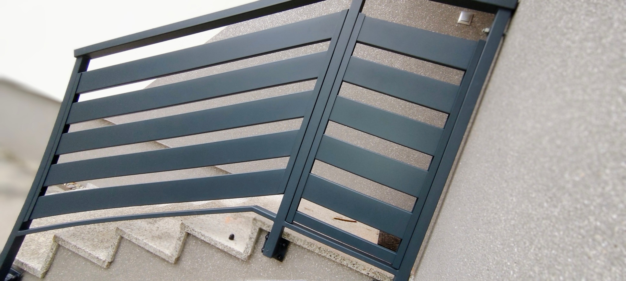 Nowoczesna, pozioma brama wjazdowa z szarych, metalowych paneli, zamontowana na schodach wejściowych. Widok z dołu, podkreślający kąt nachylenia i minimalistyczny design.