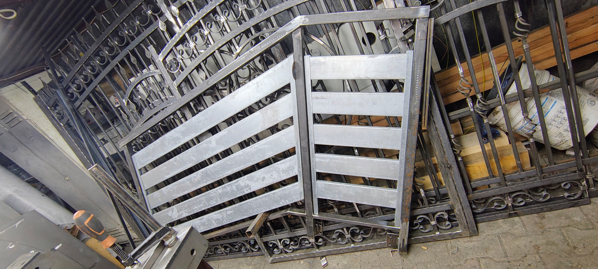 Srebrna, surowa brama z poziomymi szczeblami i ozdobne, czarne elementy bramy w tle, widoczne narzędzia ślusarskie.