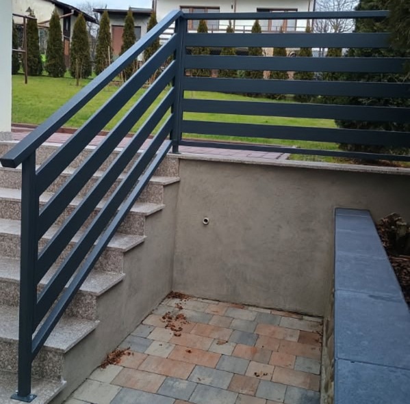 Nowoczesna, grafitowa balustrada schodowa z poziomymi szczeblami, zamontowana przy wejściu do piwnicy. Widoczne schody z jasnego kamienia i kostka brukowa.