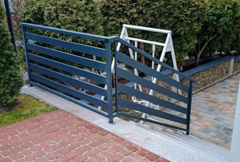 Nowoczesna, pozioma balustrada metalowa w kolorze grafitowym, zabezpieczająca schody zewnętrzne prowadzące do ogrodu. W tle widoczny huśtawka.