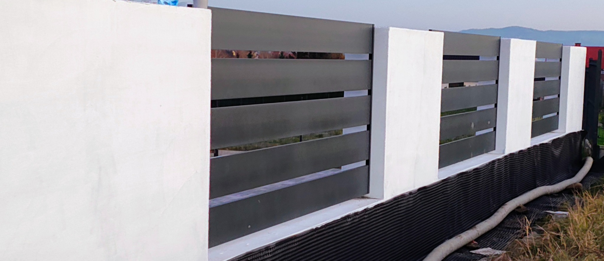 Nowoczesne ogrodzenie z poziomych, grafitowych paneli między białymi słupkami. Widoczne zabezpieczenie fundamentu. Tło stanowią wzgórza i fragment dachu.