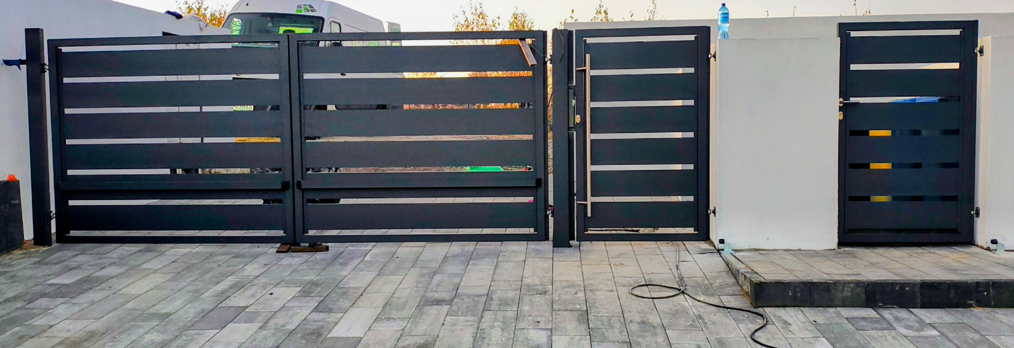 Nowoczesna, pozioma brama wjazdowa z metalu w odcieniach szarości, z widocznym mechanizmem otwierania i elementami montażowymi na tle jasnego muru.