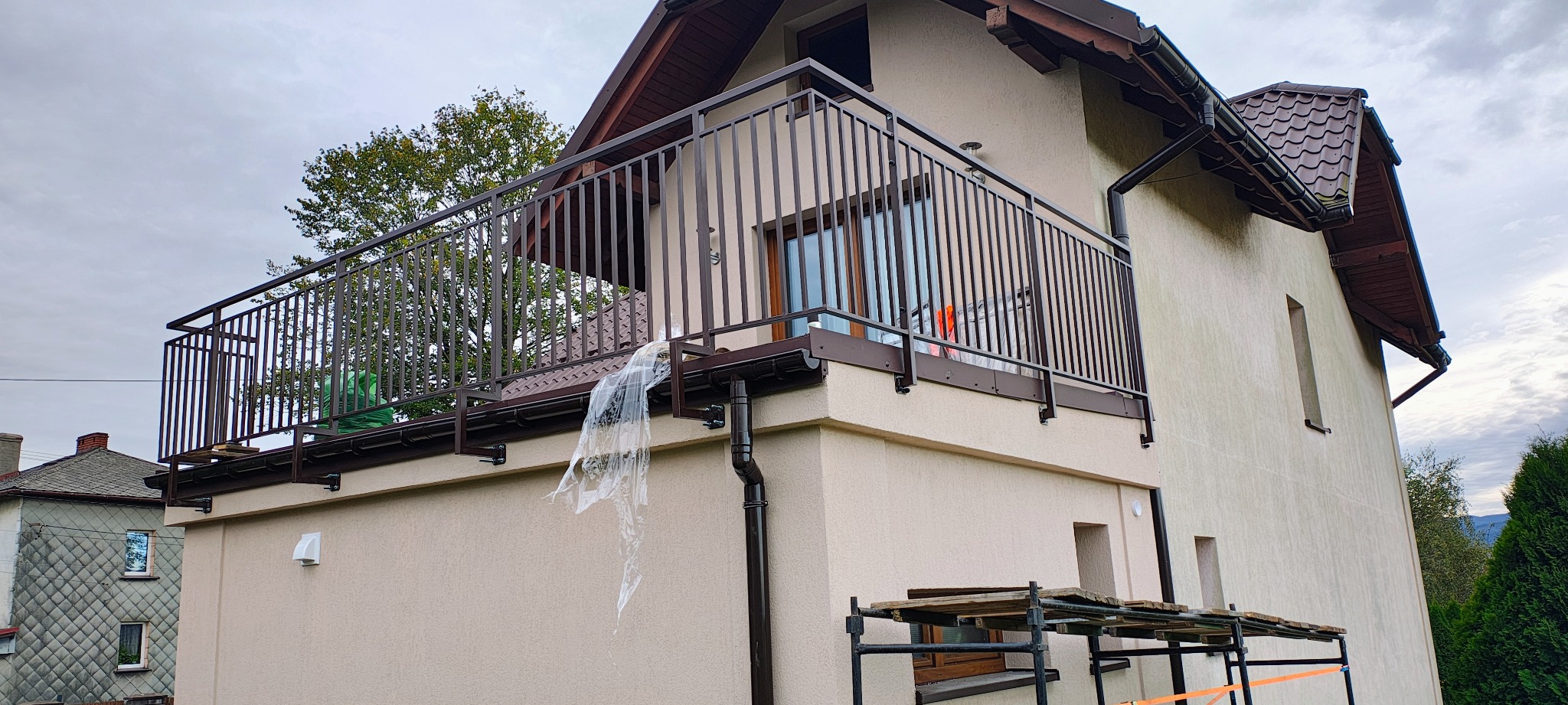 Balkon z brązową, metalową balustradą na piętrze domu z beżową elewacją. Częściowo zabezpieczony folią. W tle drzewo i budynki.