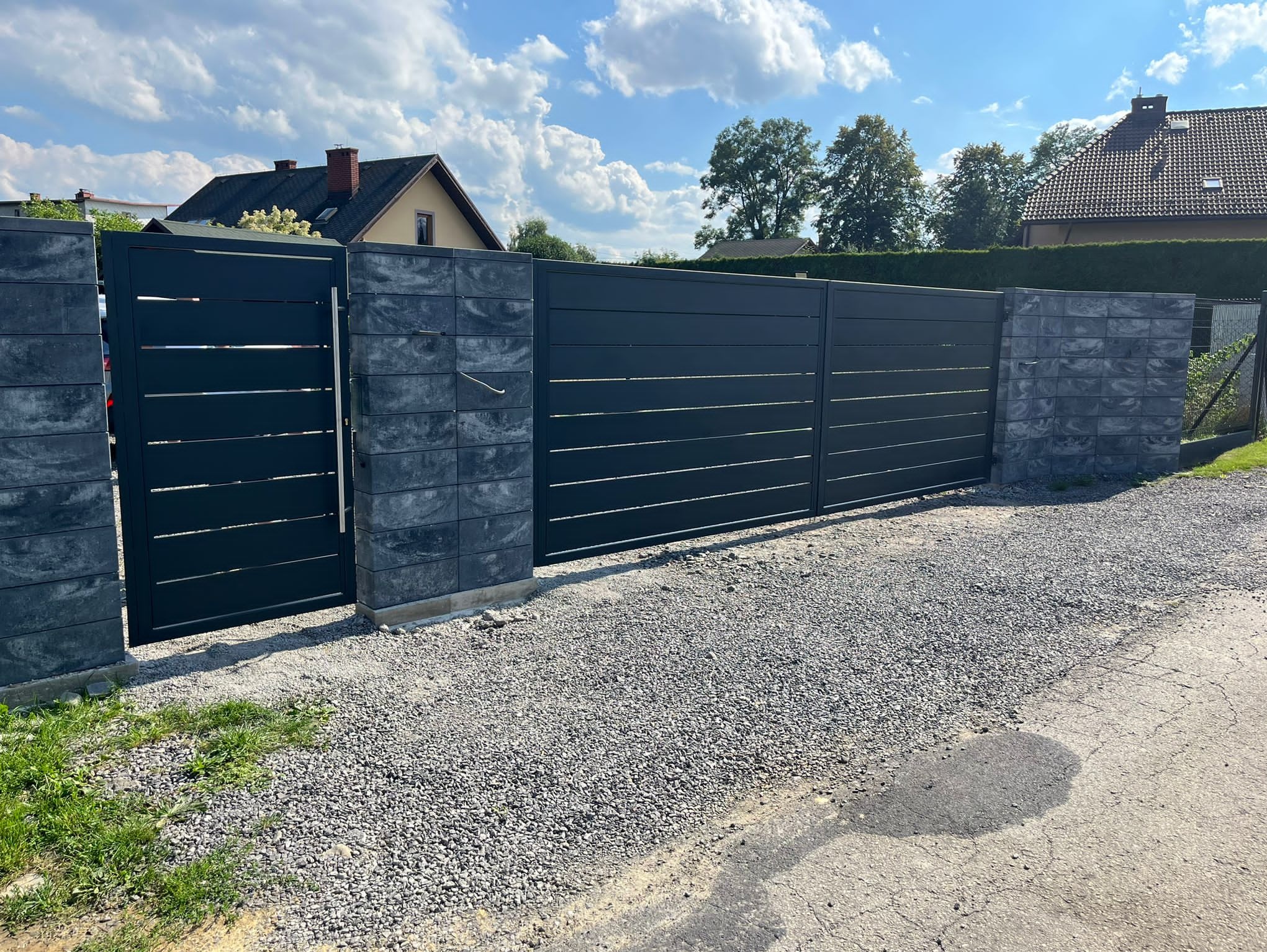 Nowoczesna brama wjazdowa z poziomymi panelami, osadzona na szarych, betonowych słupkach. Tło: dom i błękitne niebo z chmurami. Widok z zewnątrz, na żwirowym podłożu.