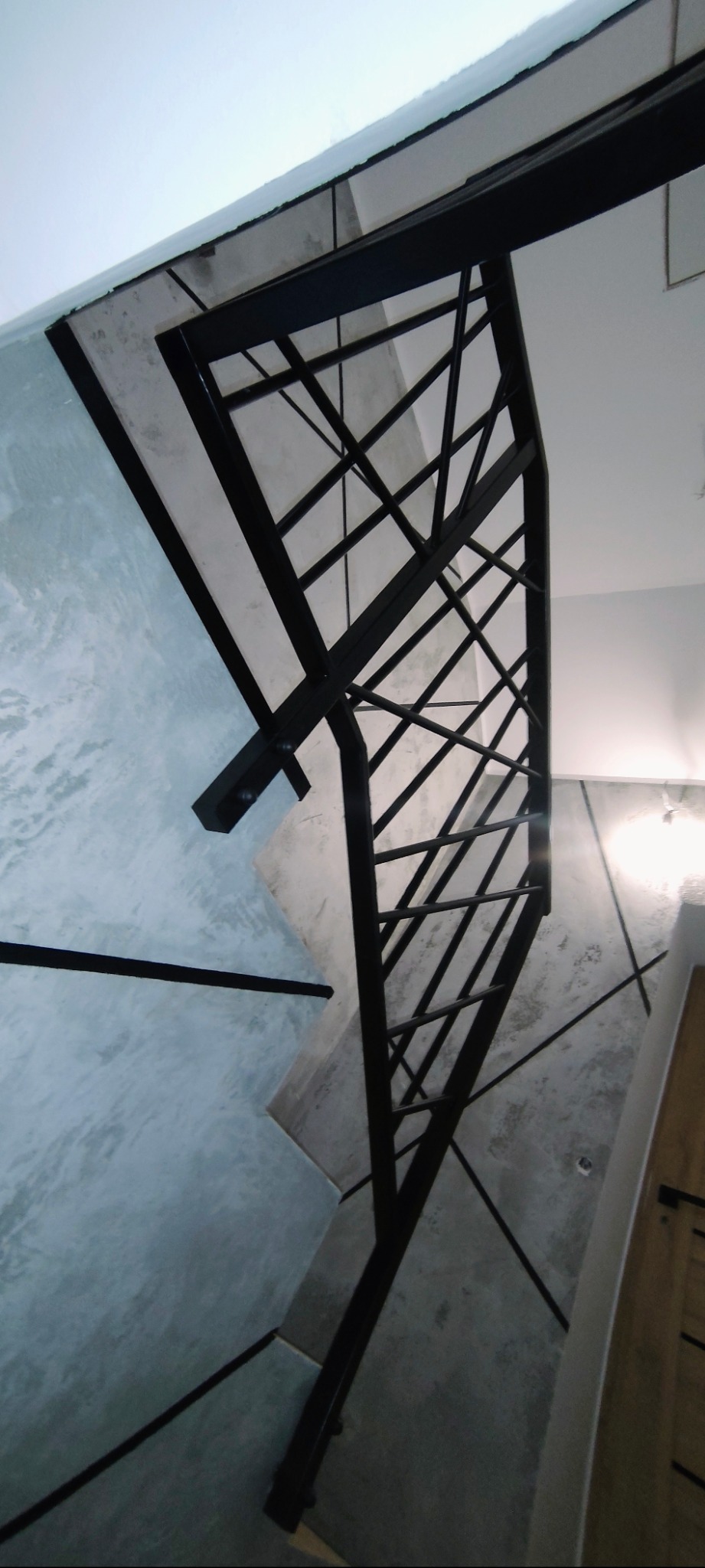 Czarna, metalowa konstrukcja schodów z geometrycznym wzorem, widziana z dołu, z fragmentem betonowej ściany o surowym wykończeniu. Minimalistyczny design.