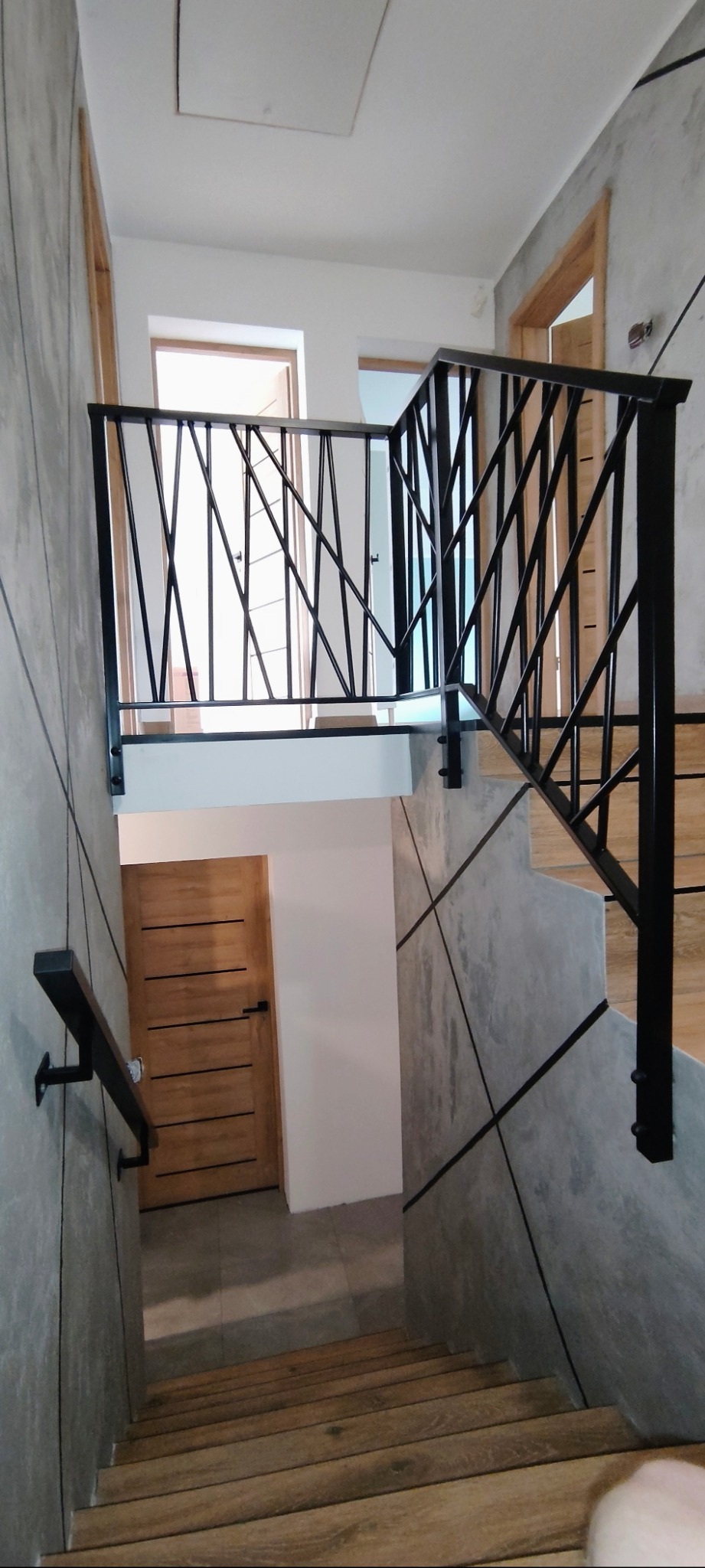 Nowoczesna balustrada schodowa z czarnymi, geometrycznymi elementami na tle betonowej ściany i drewnianych schodów. Unikalny design w loftowym wnętrzu.