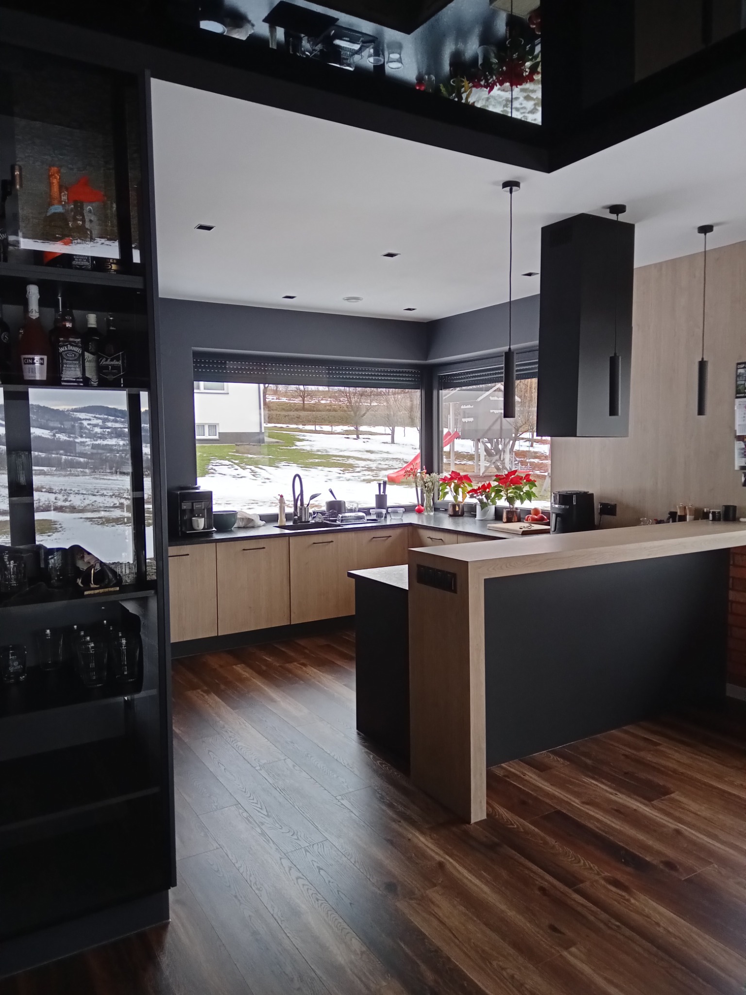 Nowoczesna kuchnia z drewnianymi frontami i czarnymi akcentami. Duże okno z widokiem na zimowy krajobraz. Na blacie kwiaty w doniczkach.