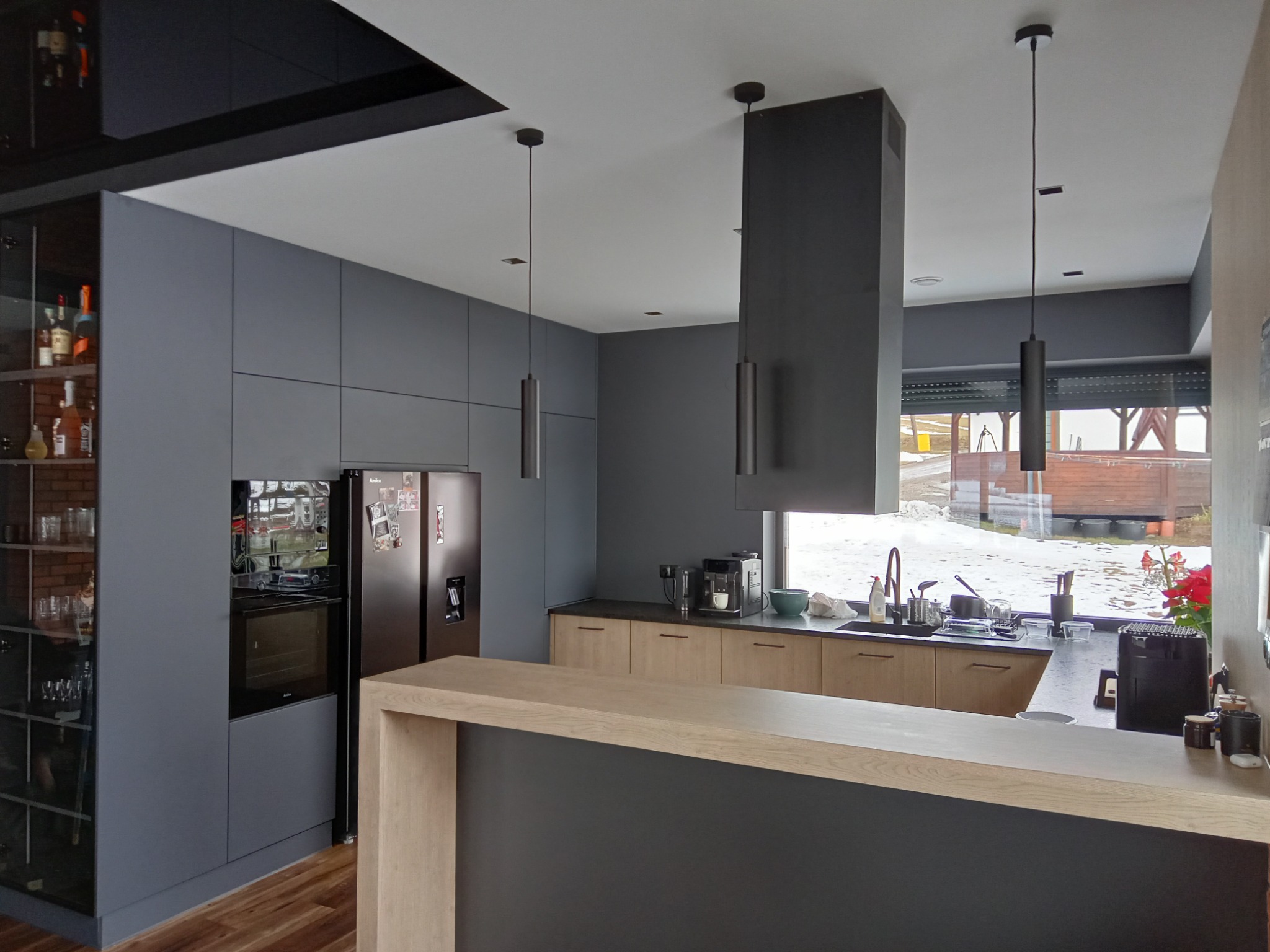 Nowoczesna kuchnia z szarymi frontami i drewnianym blatem, wyspą. Widoczne oświetlenie punktowe i przeszklona witryna z barkiem. Za oknem zimowy krajobraz.