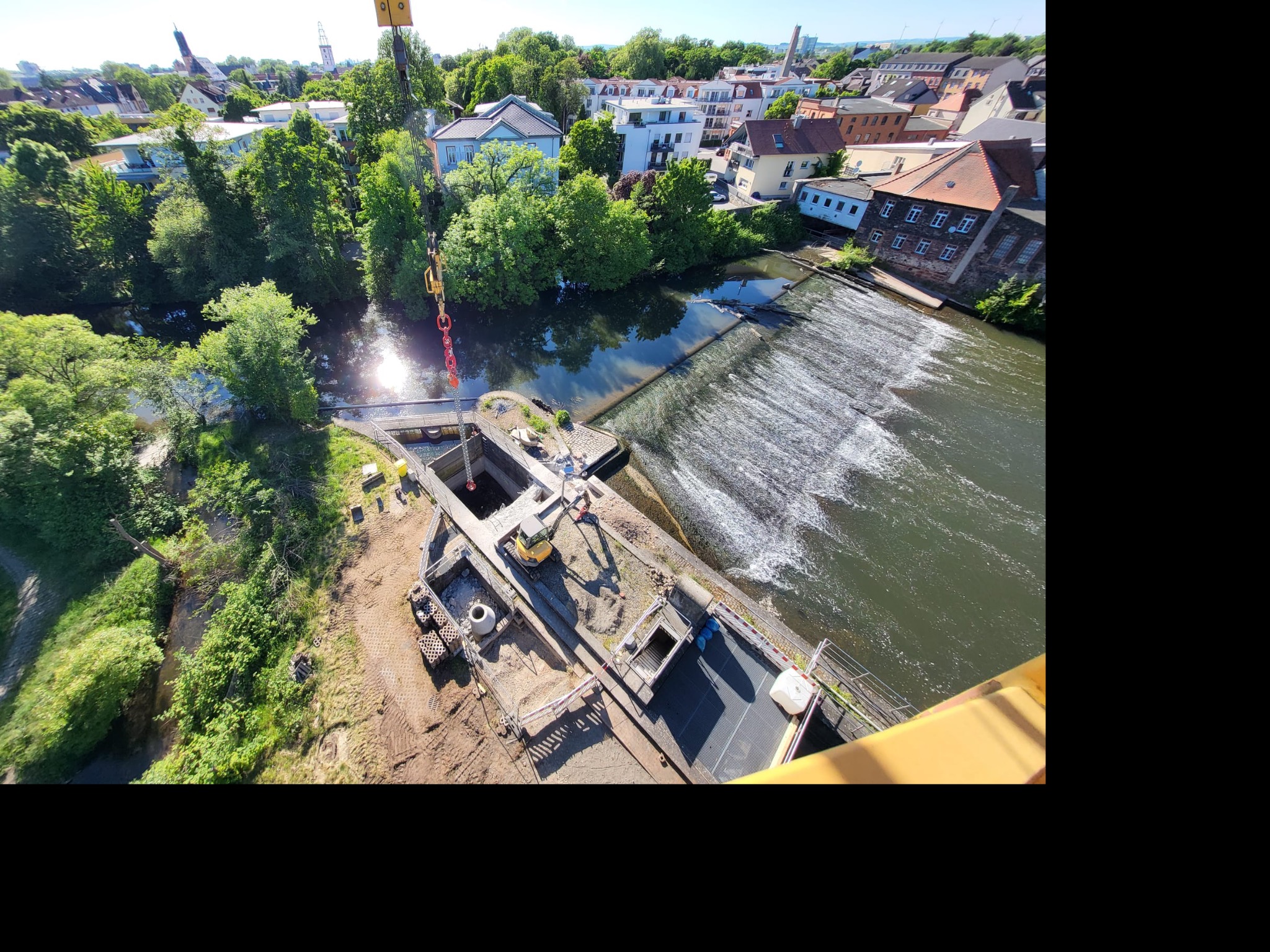 Prace ziemne przy rzece z użyciem dźwigu i koparki. Widok z góry na plac budowy, gdzie wykonywane są wykopy i prace związane z fundamentami, w tle zabudowa miejska.
