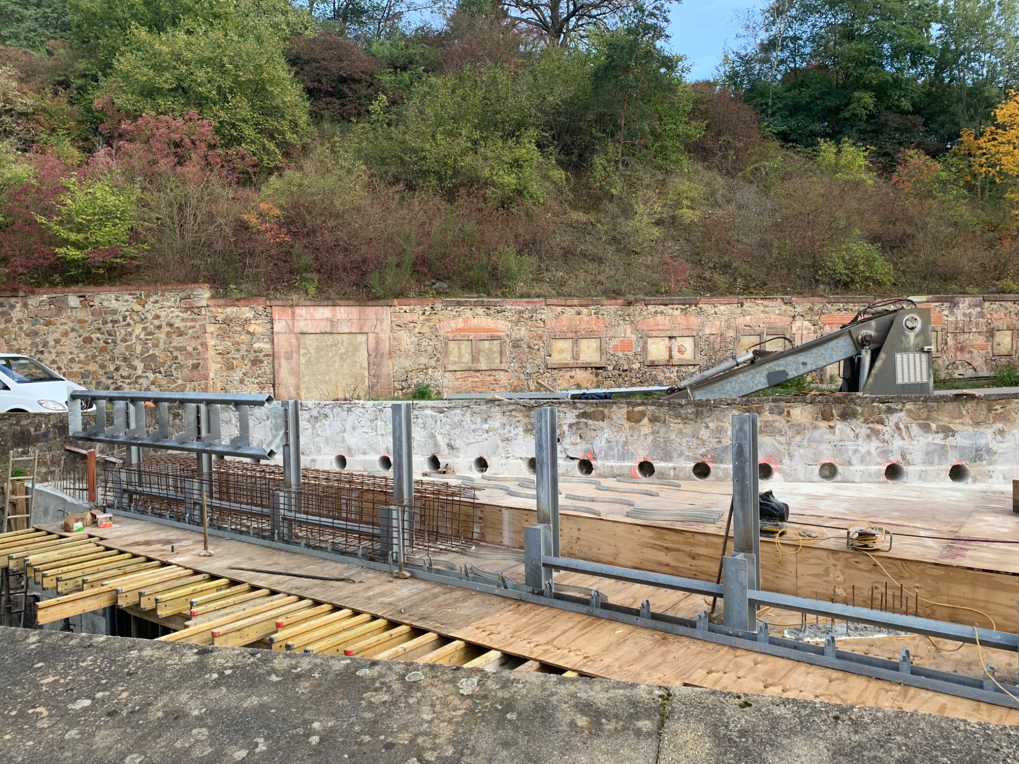 Etap budowy: szalunki i zbrojenie fundamentów na tle kamiennej ściany i zieleni. Widoczne drewniane elementy konstrukcyjne i stalowe pręty zbrojeniowe.