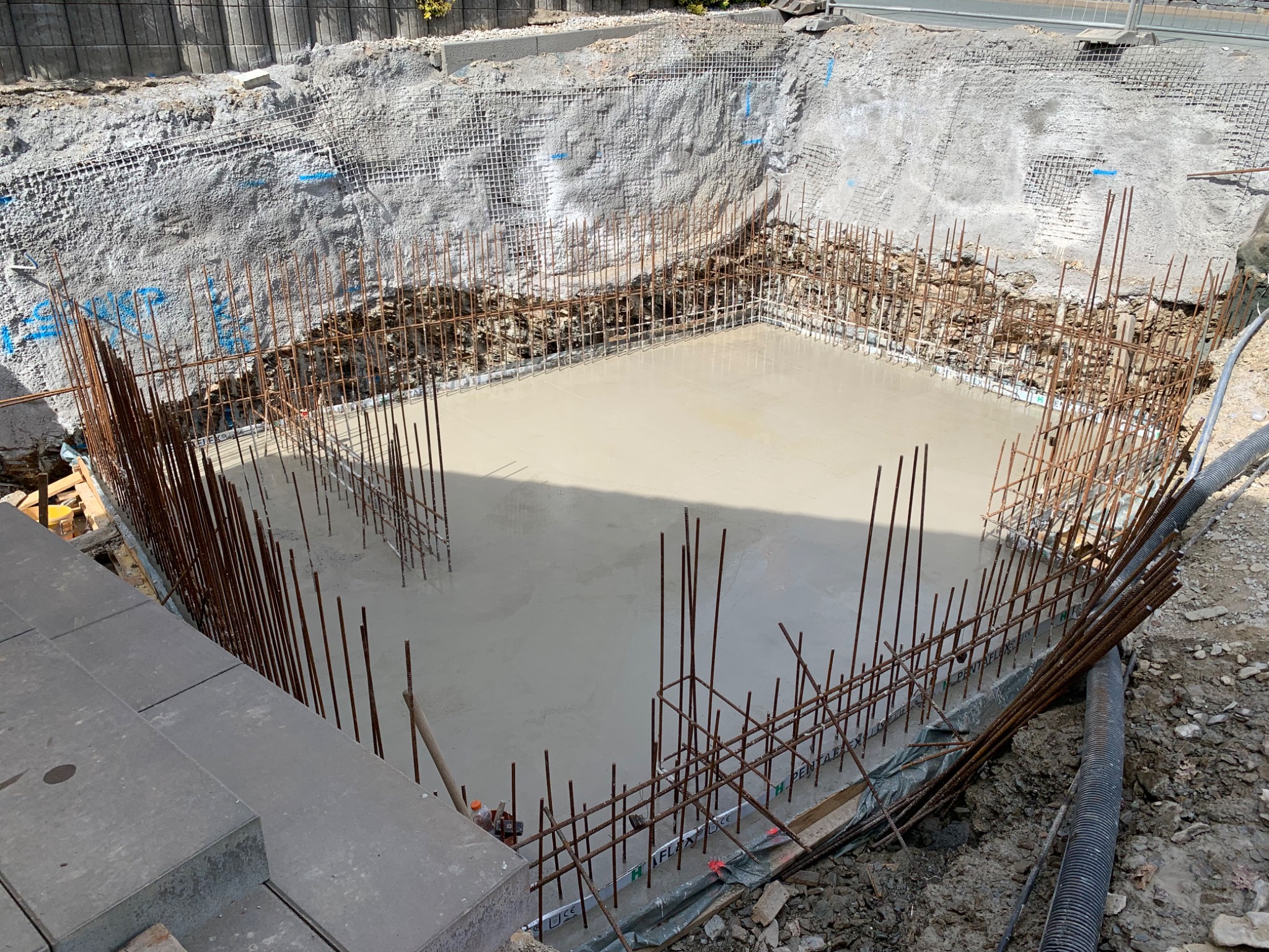 Zalany betonem fundament, zbrojenie wystaje ponad powierzchnię. Widoczne zabezpieczenie wykopu siatką. Świeży beton w wykopie pod budowę.