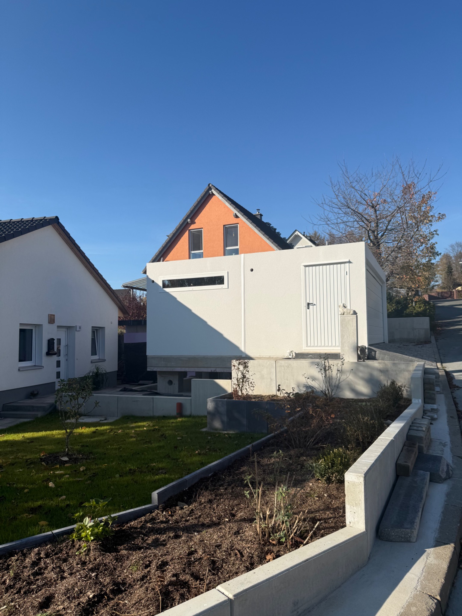 Nowoczesny, biały budynek z płaskim dachem, częściowo pod ziemią, z białymi drzwiami i oknem, otoczony zielenią i elementami betonowymi, na tle domów i błękitnego nieba.