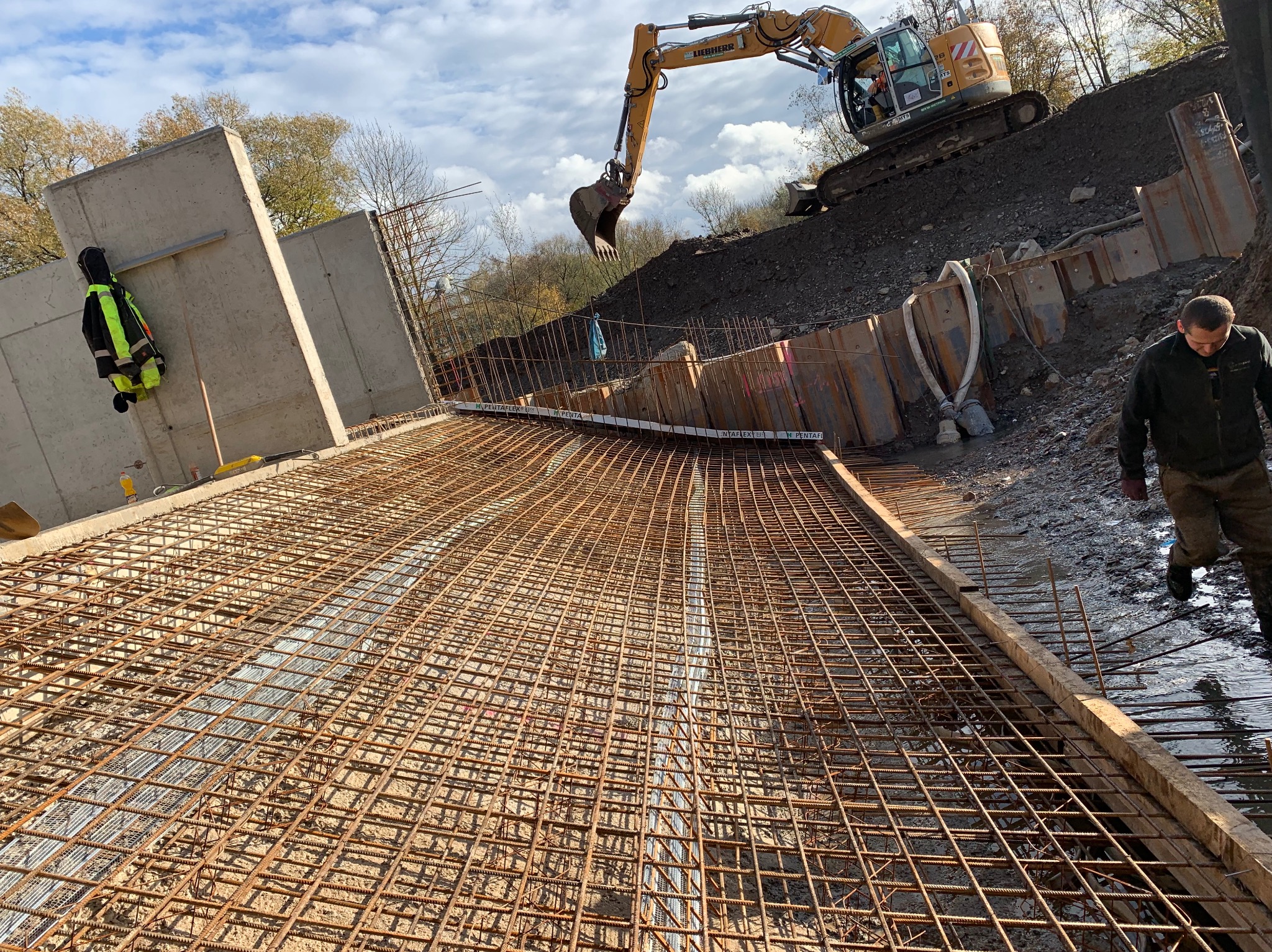 Szeroki kadr na zbrojenie fundamentów z wykopem i koparką Liebherr w tle. Na pierwszym planie siatka zbrojeniowa, a w oddali pracownik budowlany i betonowe ściany.