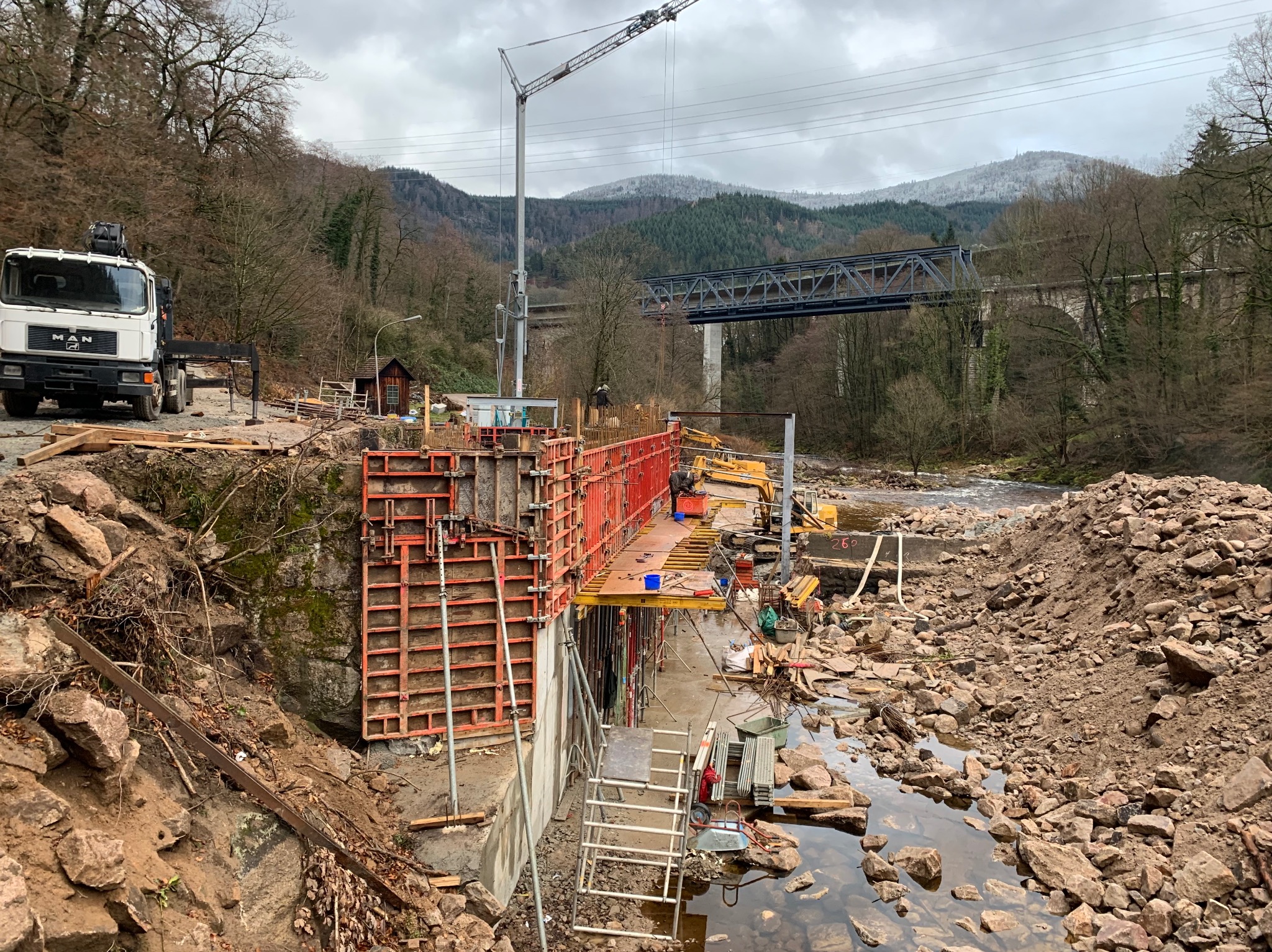 Budowa mostu: szalunek, dźwig, i ciężarówka MAN na tle gór i lasów. Widoczne elementy konstrukcyjne i sprzęt budowlany. Etap wylewania fundamentów.