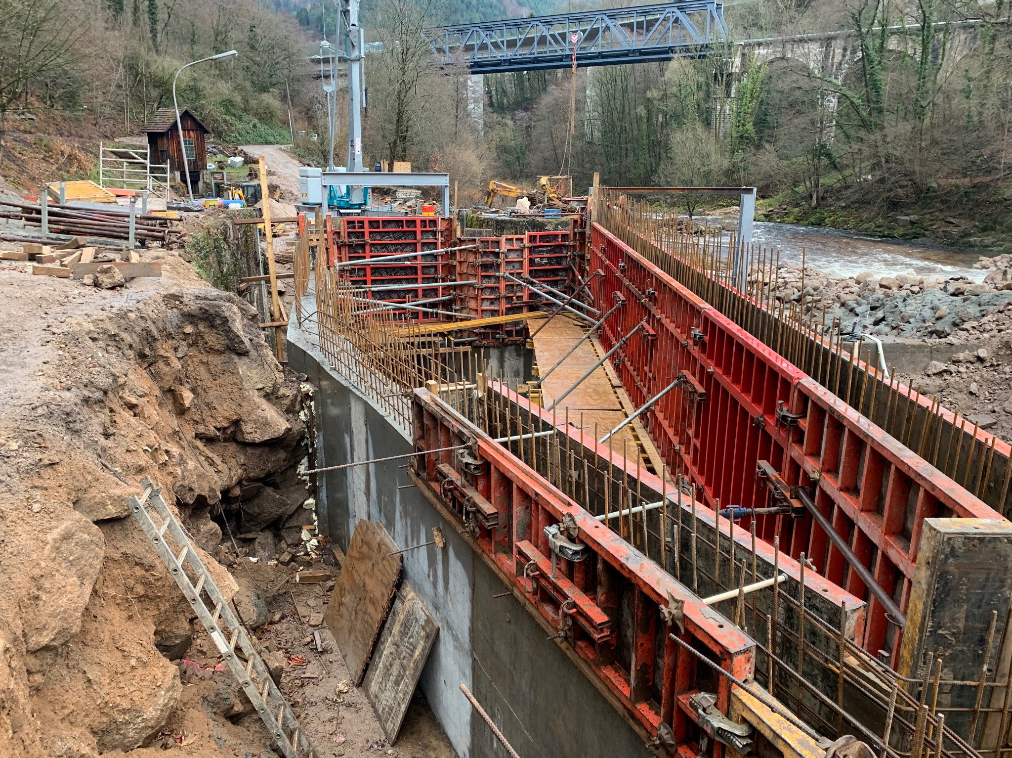 Budowa żelbetowej konstrukcji przy rzece z widocznymi szalunkami i zbrojeniami. W tle most i zalesione wzgórza. Widoczna drabina i wykop ziemny.