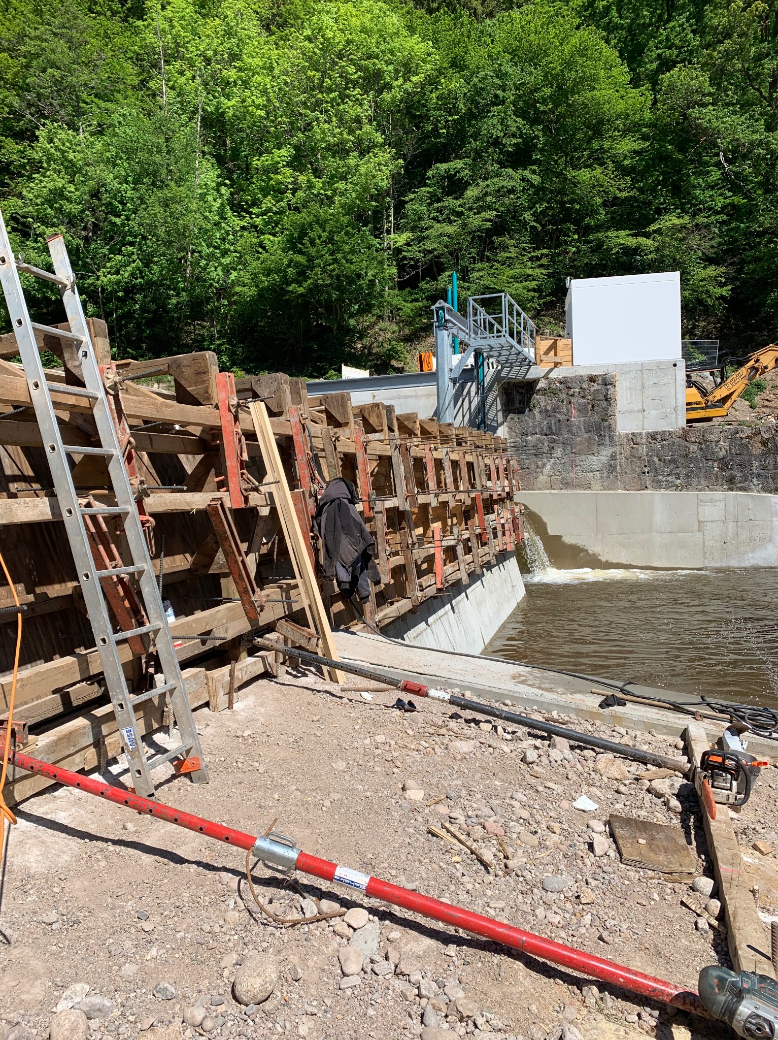 Budowa zapory wodnej: drewniane szalunki, betonowa konstrukcja, w tle las. Widoczny sprzęt budowlany i narzędzia. Woda przelewa się przez konstrukcję.