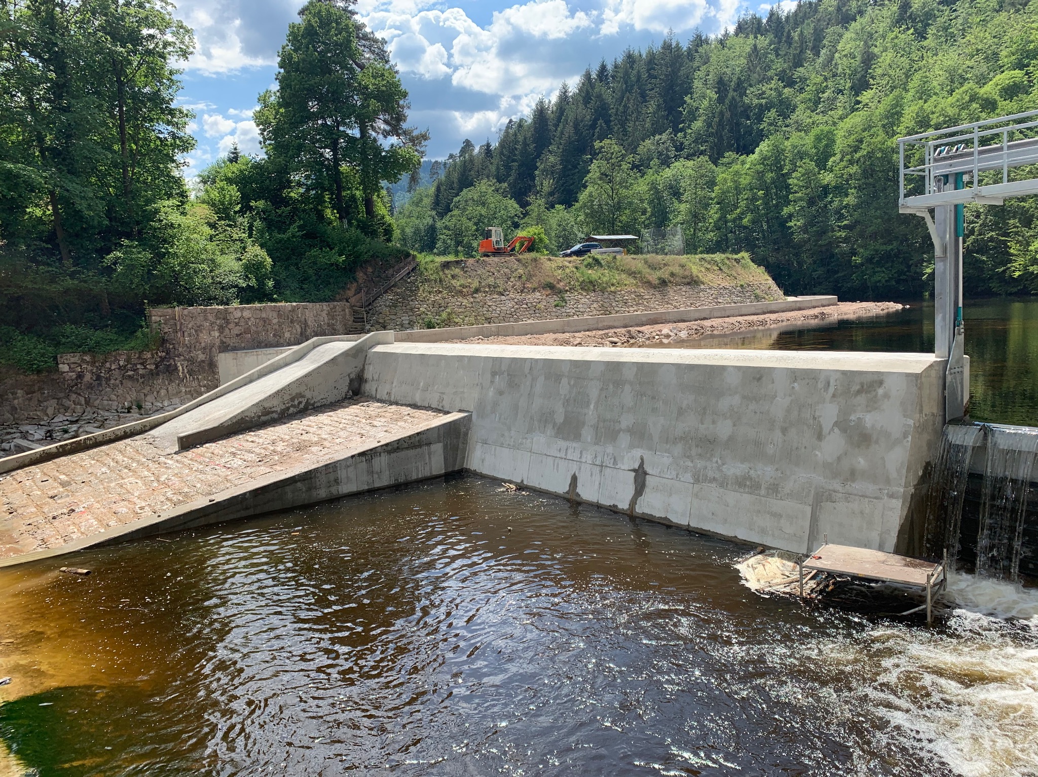 Betonowa konstrukcja hydrotechniczna z przelewem, w tle zalesione wzgórze i sprzęt budowlany. Ujęcie w słoneczny dzień.