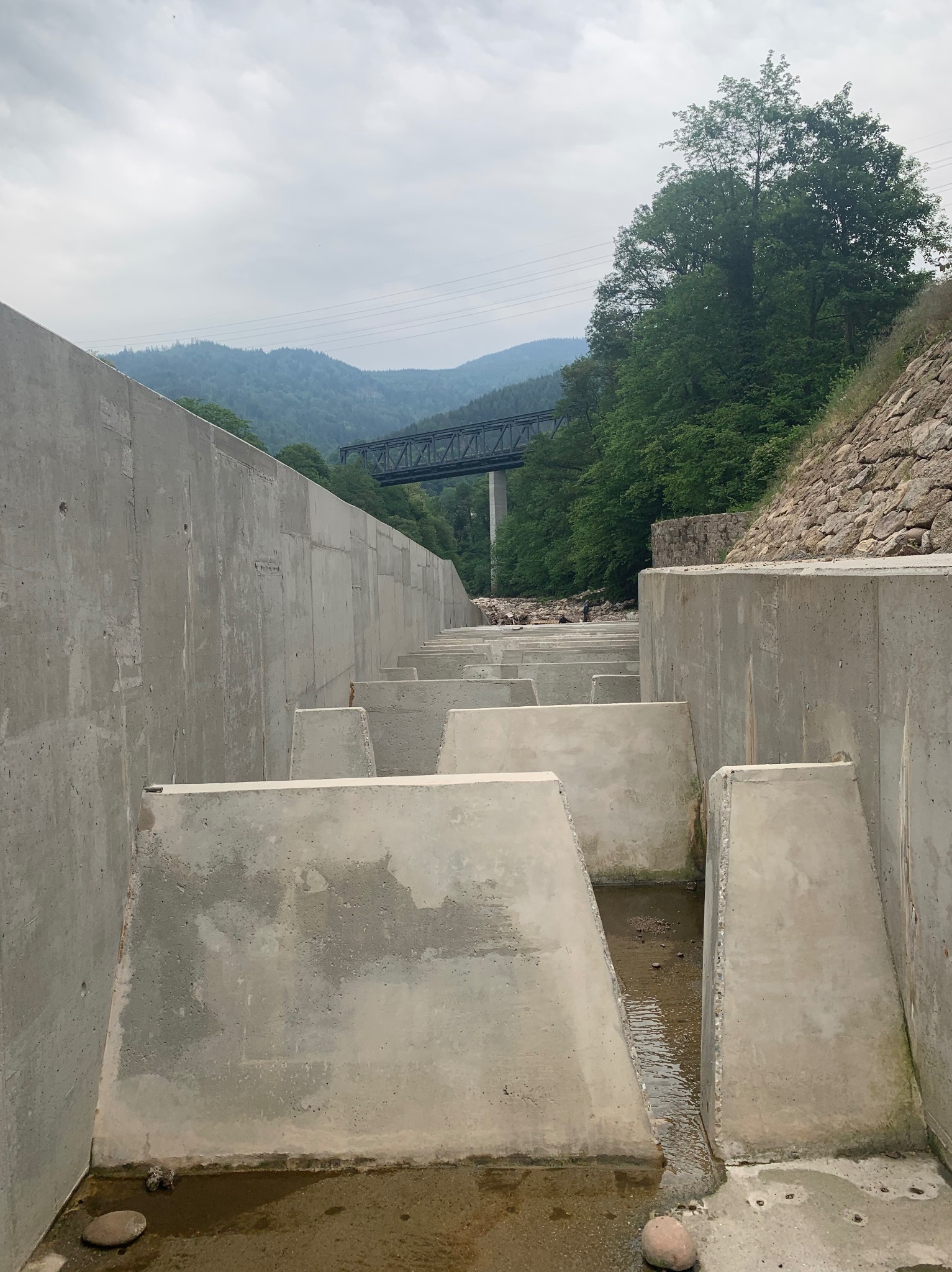 Betonowe kaskady na rzece z widokiem na góry i most. Konstrukcja hydrotechniczna z surowego betonu w otoczeniu zieleni i naturalnego krajobrazu.