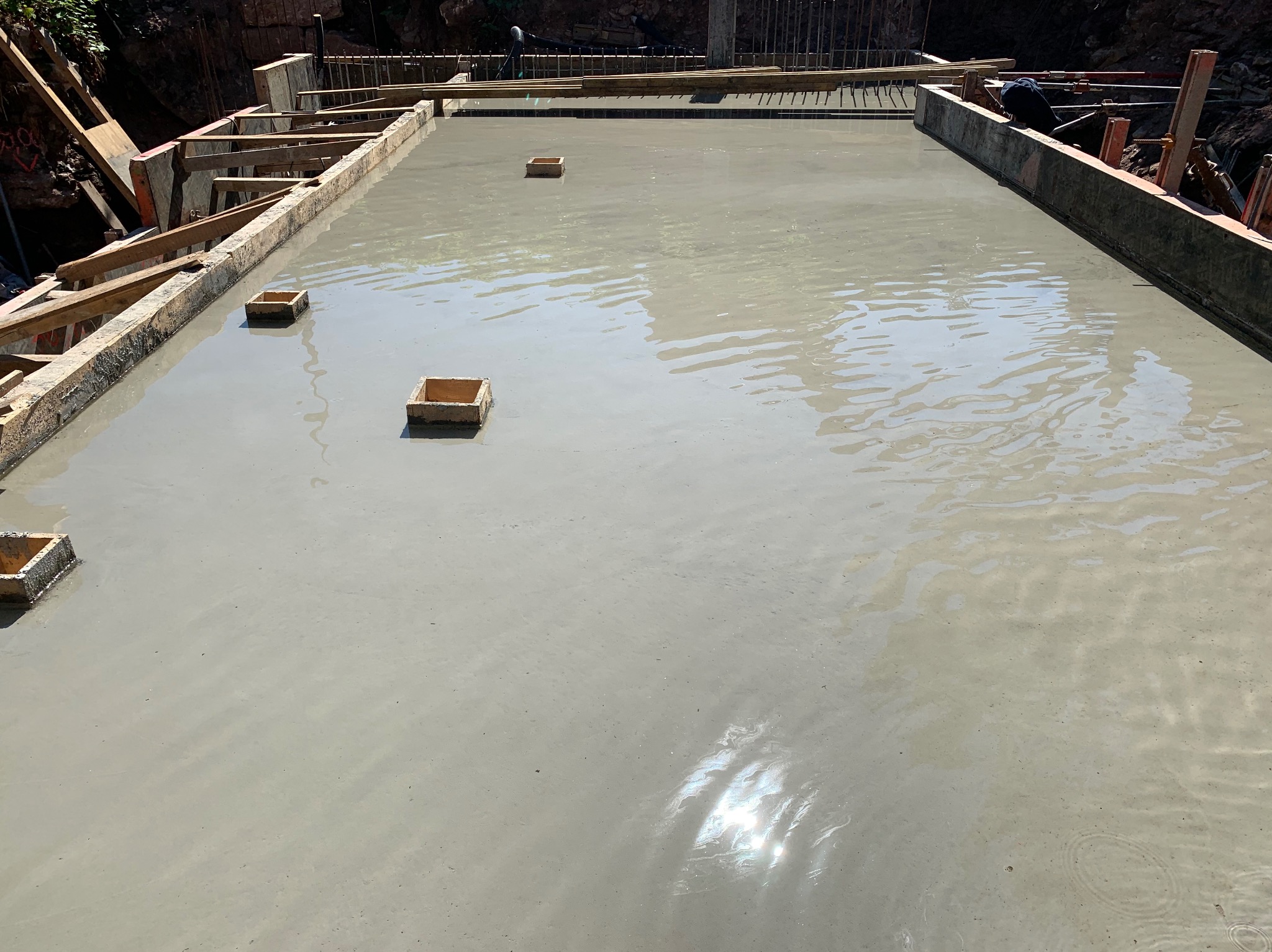 Zalany fundament z drewnianym szalunkiem i wystającymi elementami dystansowymi. Widoczna zbrojona konstrukcja w tle. Etap budowy.