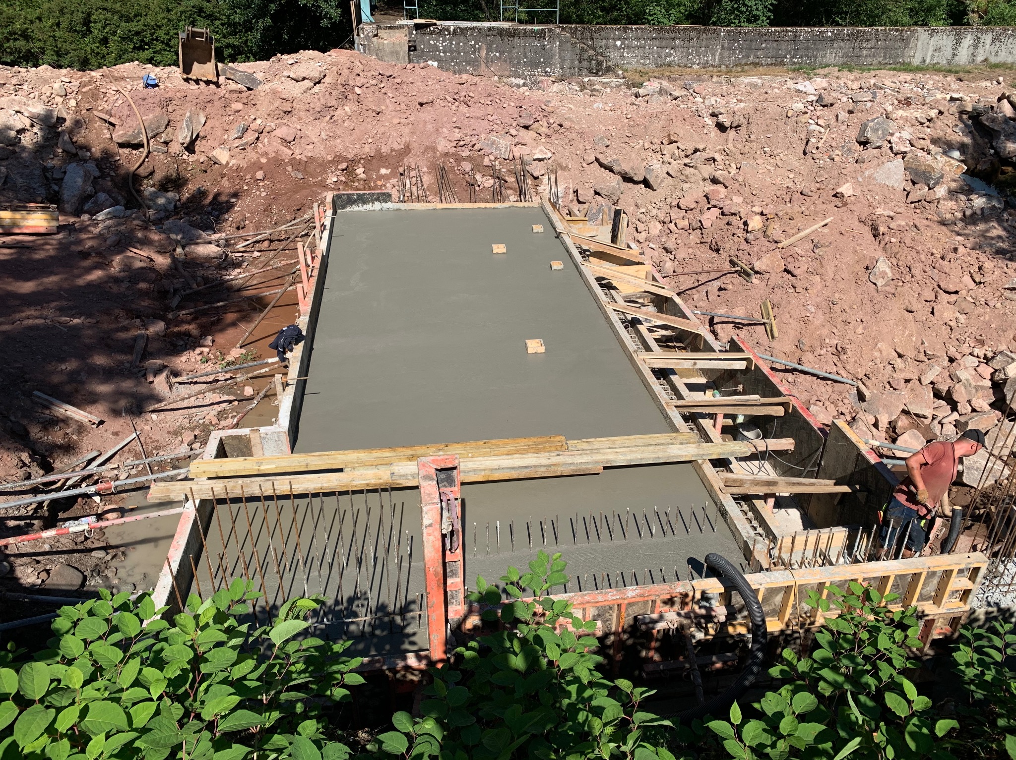 Wylany beton w szalunku z widocznym zbrojeniem. W tle ziemia i gruz, na pierwszym planie zieleń. Prace budowlane w toku, betonowa konstrukcja.