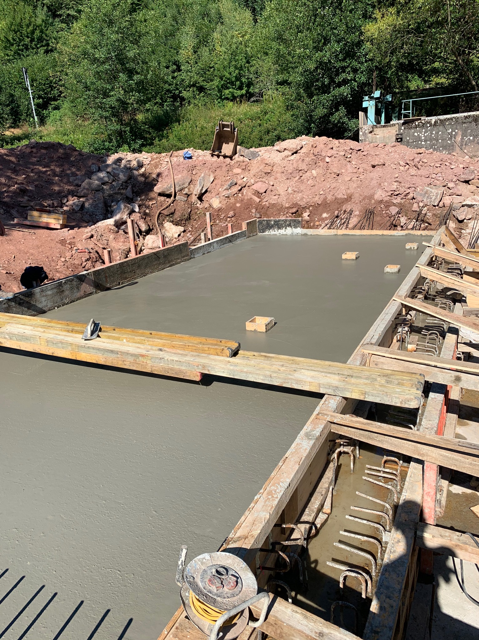 Wylany fundament z drewnianym szalunkiem i zbrojeniem, na tle wykopu ziemnego. Widoczne narzędzia i akcesoria budowlane. Górny kadr ukazuje zieleń.