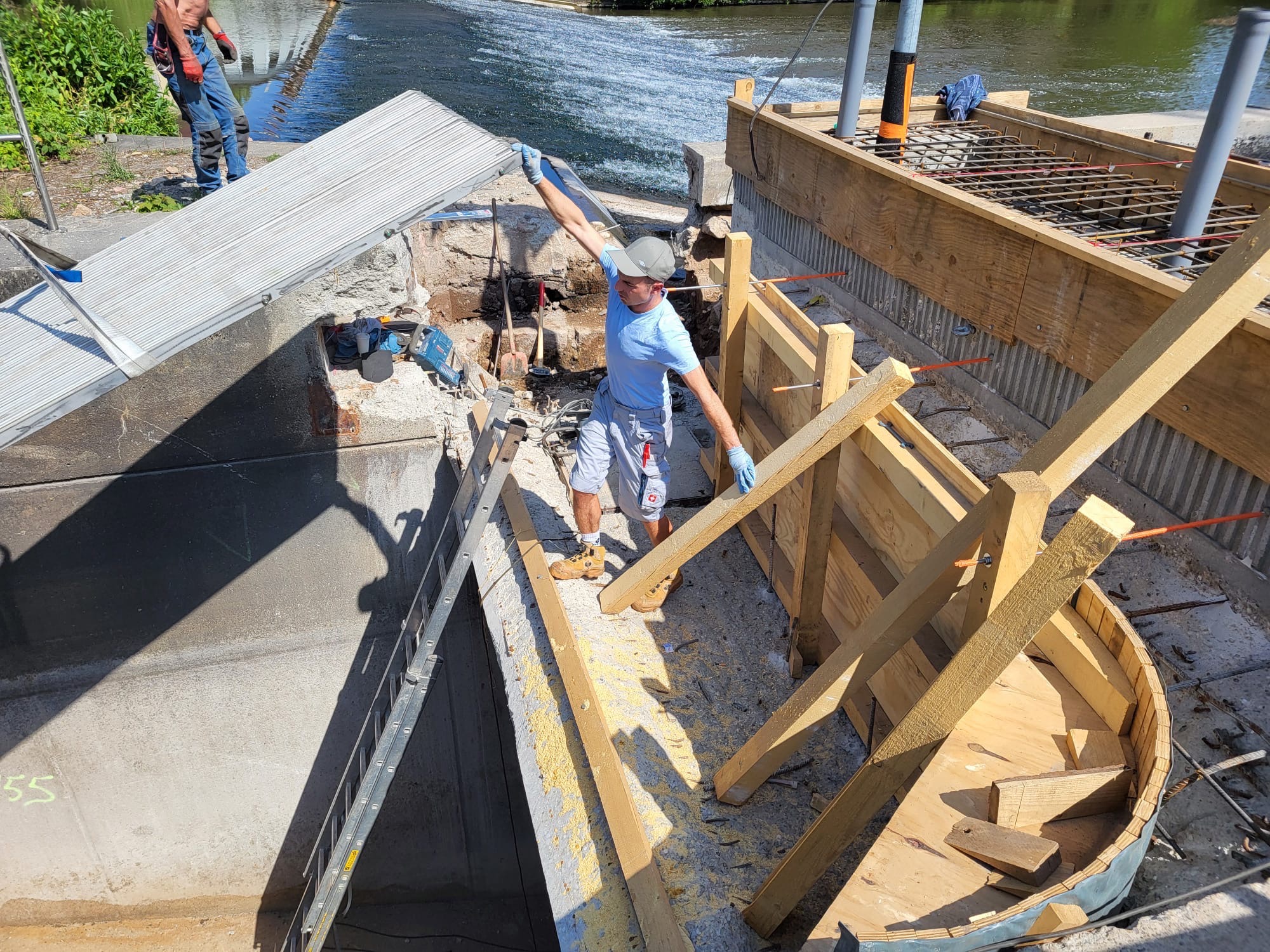 Prace przy budowie zbrojonej konstrukcji betonowej nad wodą. Widoczny pracownik w trakcie montażu szalunków i zbrojenia. W tle woda i roślinność.