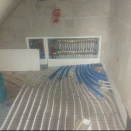 HYDROMAR - Rozłożone białe rury ogrzewania podłogowego na siatce, z niebieskimi elementami mocującymi. W tle rozdzielacz w białej obudowie na szarej ścianie.